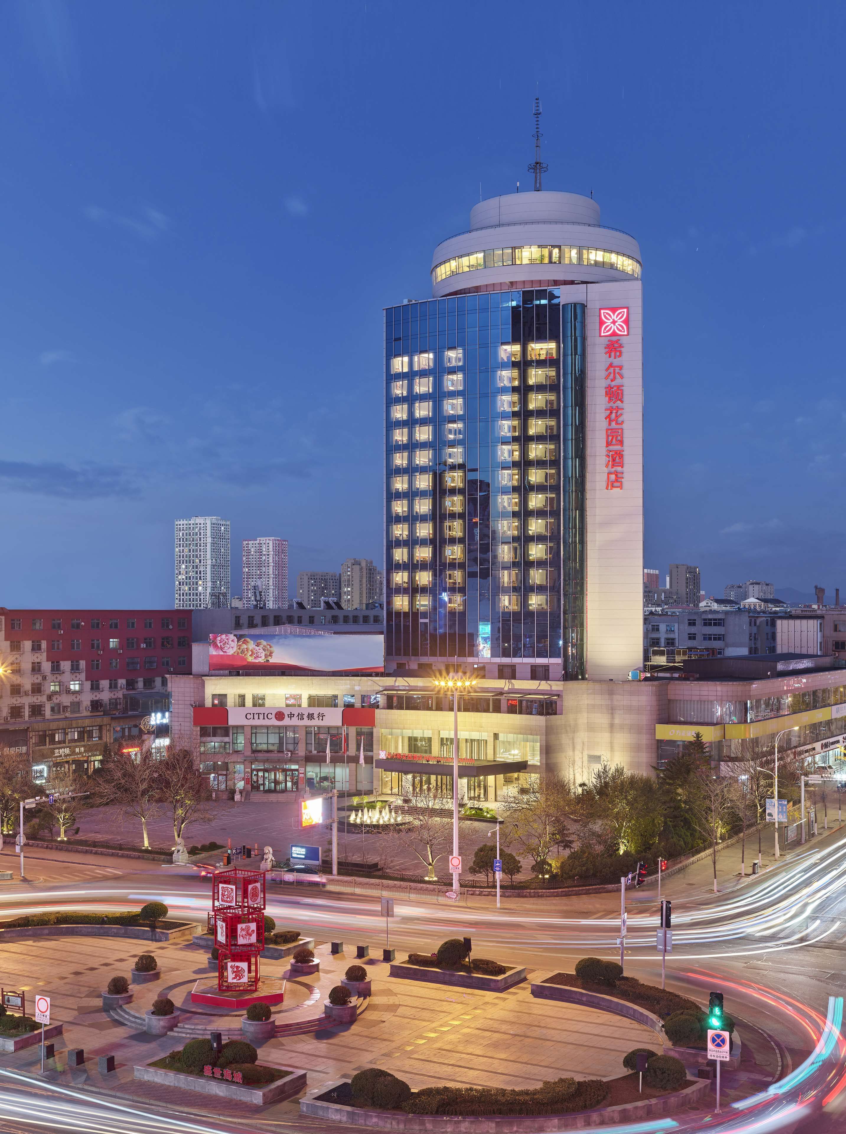 Een modern hotel met een cilindrische toren, omgeven door stadsgebouwen en een goed verlicht plein met geometrisch landschap.