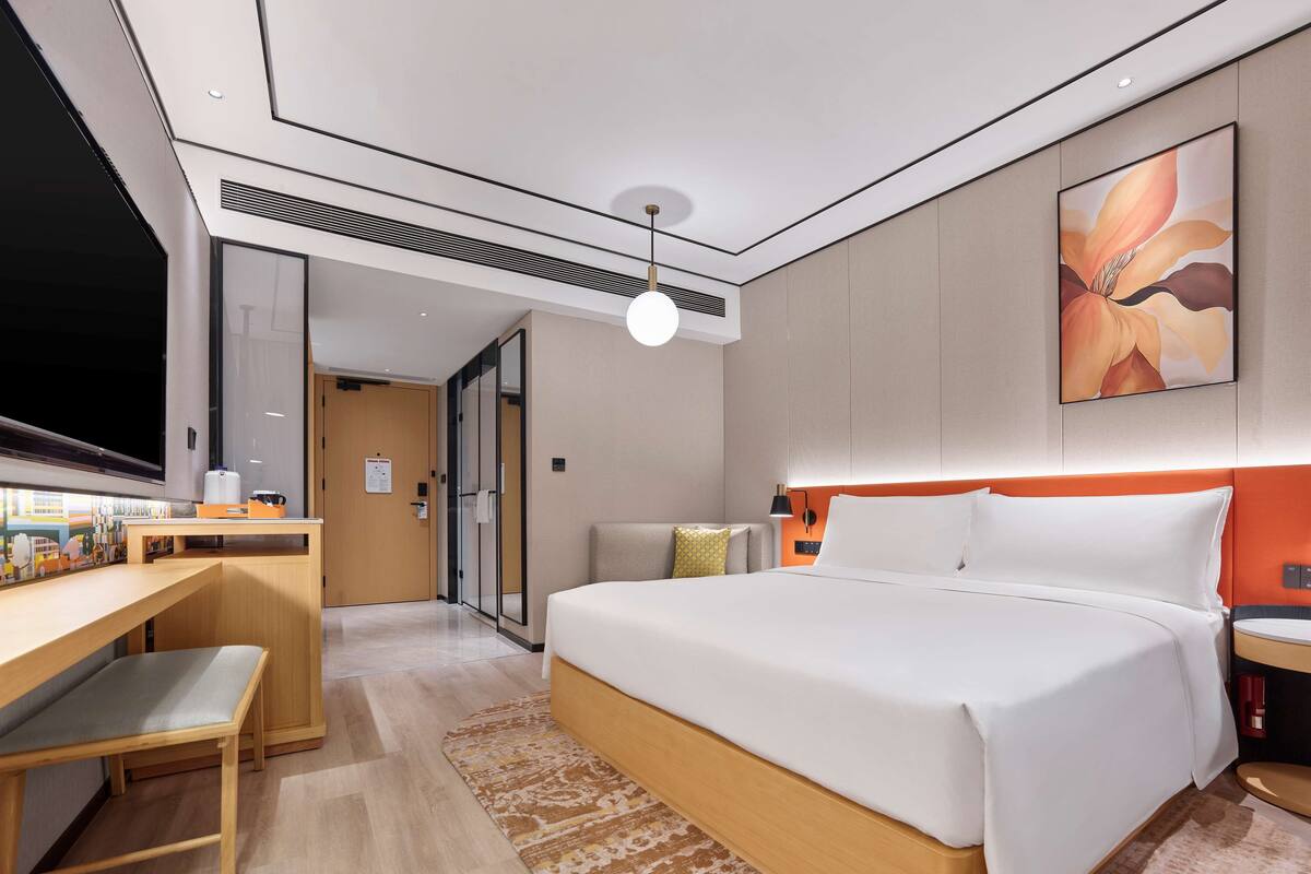 A modern hotel room with a large bed, a desk with a chair, a TV, and a painting on the wall.