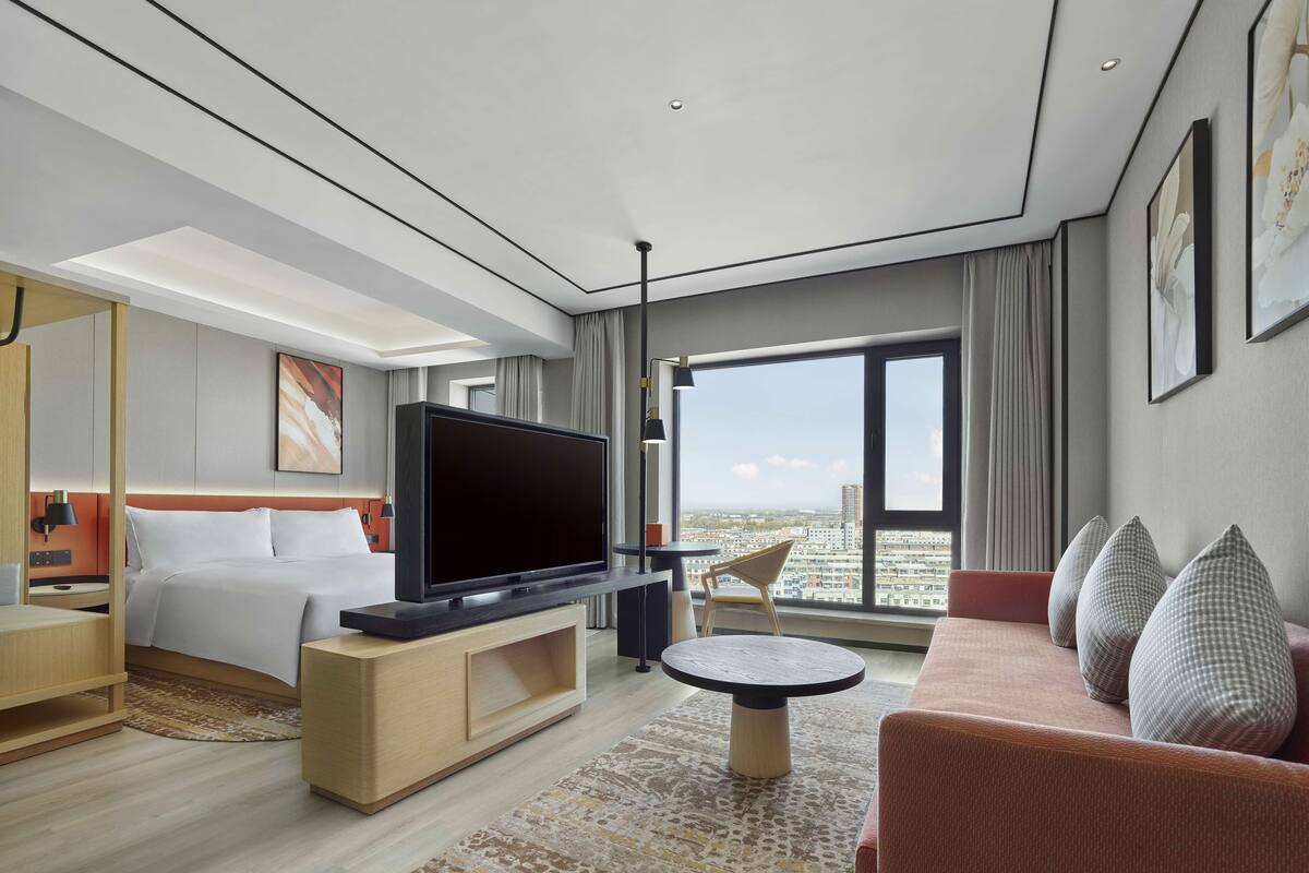 A modern hotel room with a large bed, a flat-screen TV, a sofa, and a dining table.