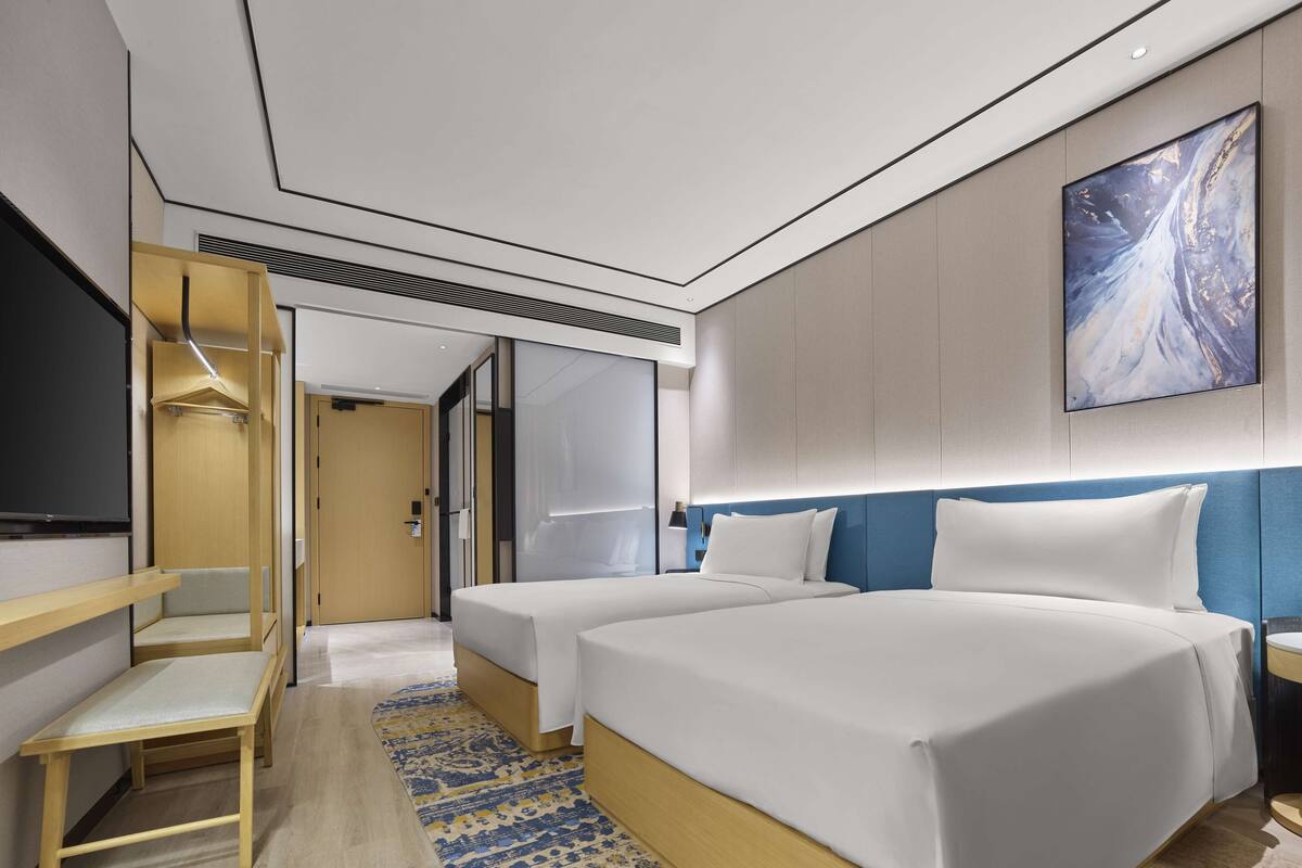 A modern hotel room with two beds, a large mirror, a painting, and a wooden bench.