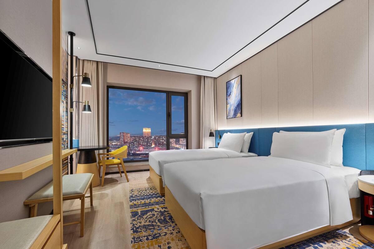 A hotel room with two beds, a large window with a city view, a flat-screen TV, and a small seating area.