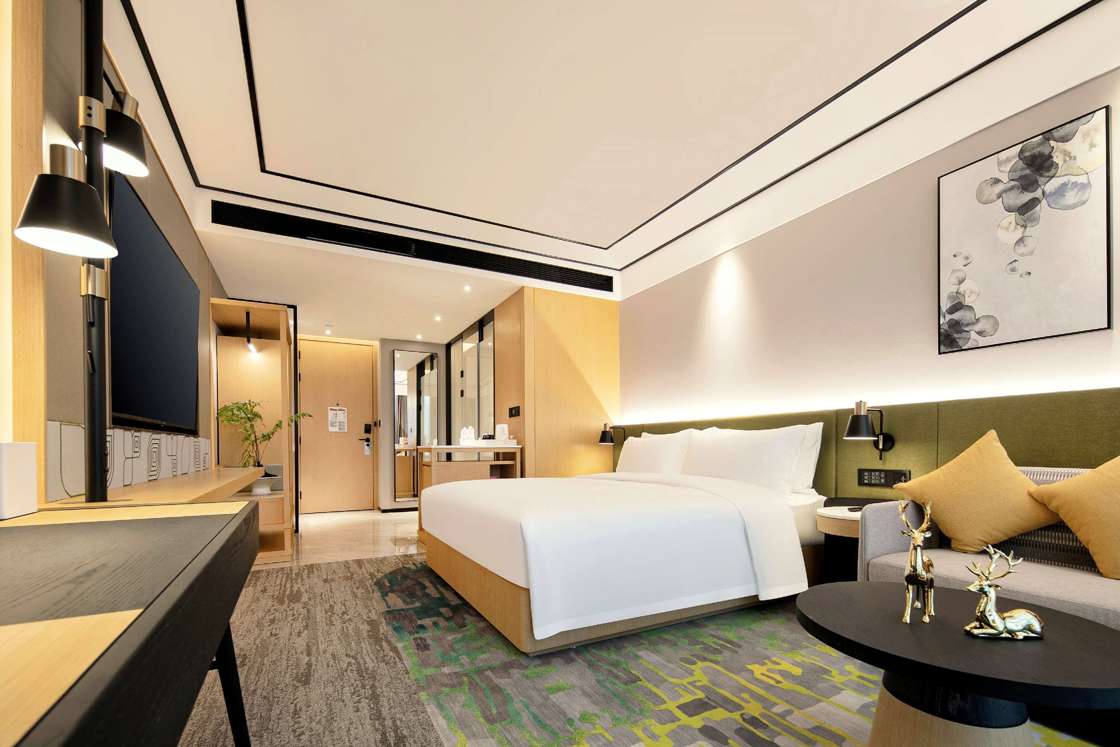 A modern hotel room with a large bed, a desk with a flat-screen TV, a sofa, and a coffee table.