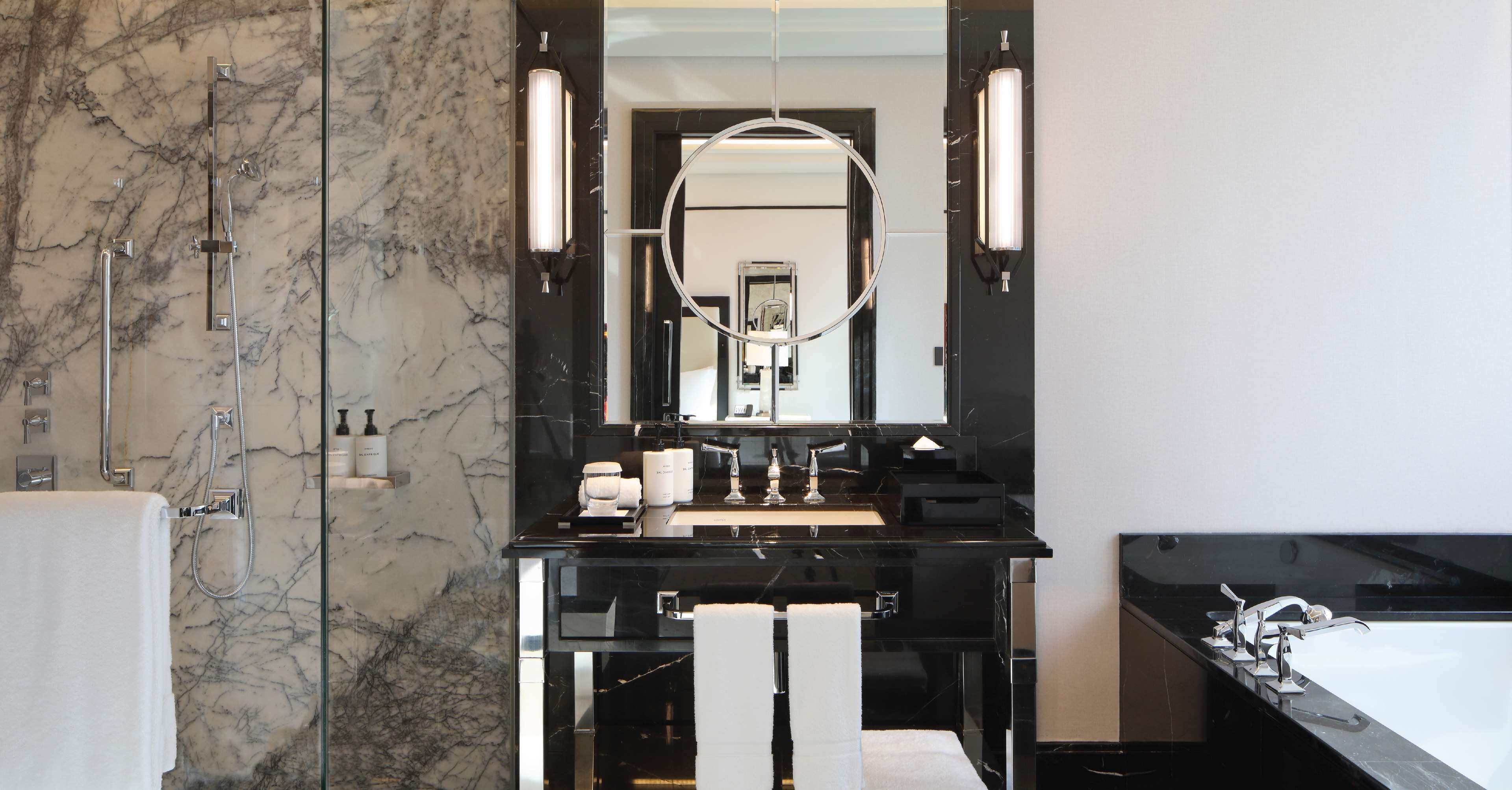 A modern bathroom with a marble wall, a black countertop, a large mirror, and a separate bathtub area.