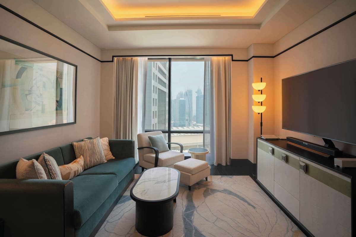Une chambre d’hôtel moderne équipée d’un canapé, d’un fauteuil, d’un pouf, d’une table basse, d’une télévision et d’une grande fenêtre donnant sur la ville.