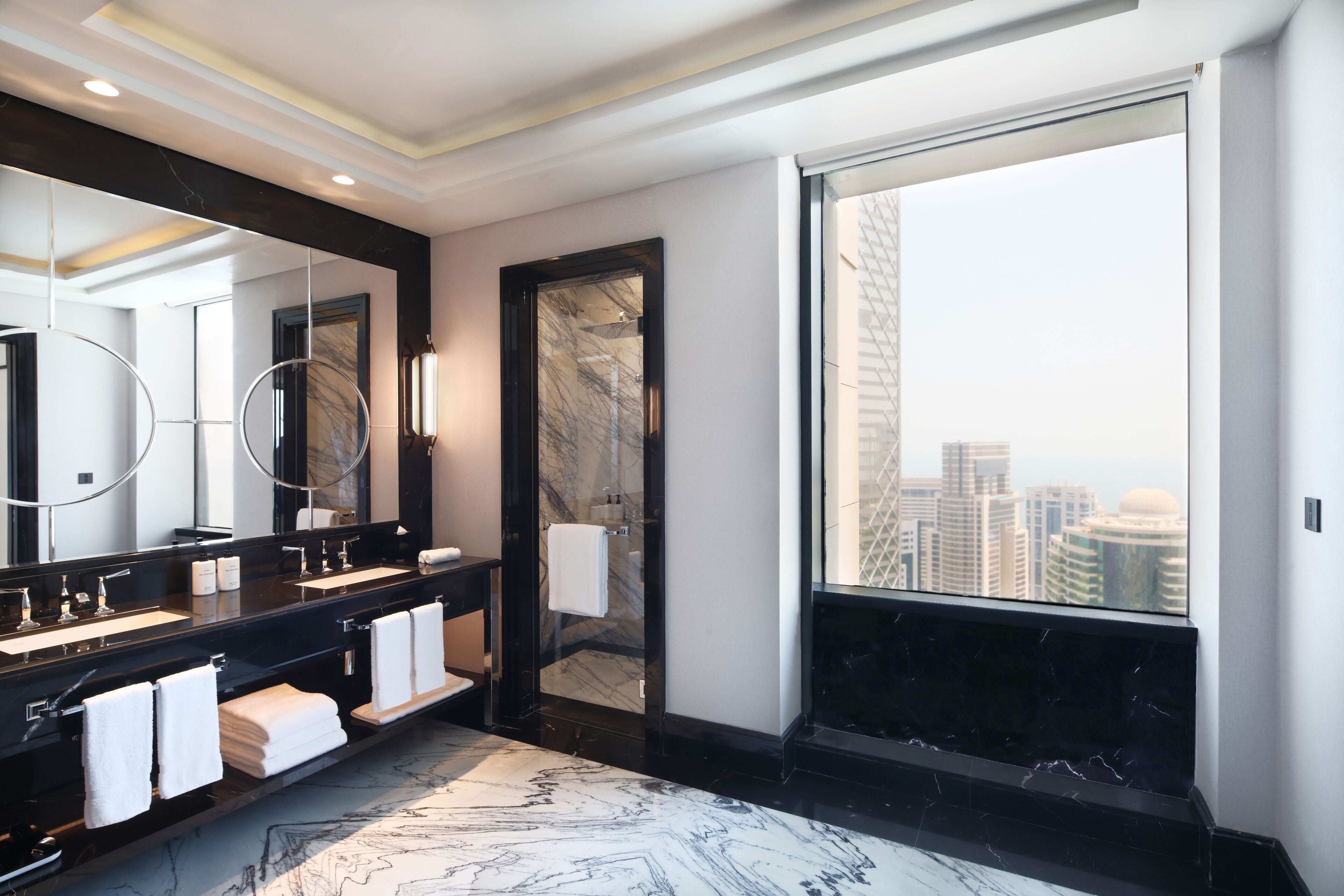 Un baño moderno con un espejo grande, lavamanos dobles y vistas a la ciudad a través de un ventanal amplio.