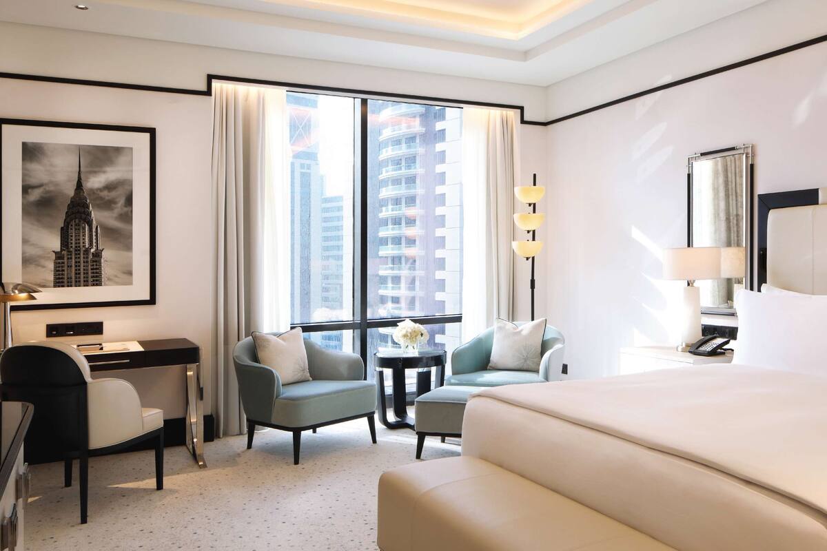 Une chambre d’hôtel avec un grand lit, un bureau avec une chaise, deux fauteuils, une petite table avec un vase, une lampe et une grande fenêtre avec des rideaux.