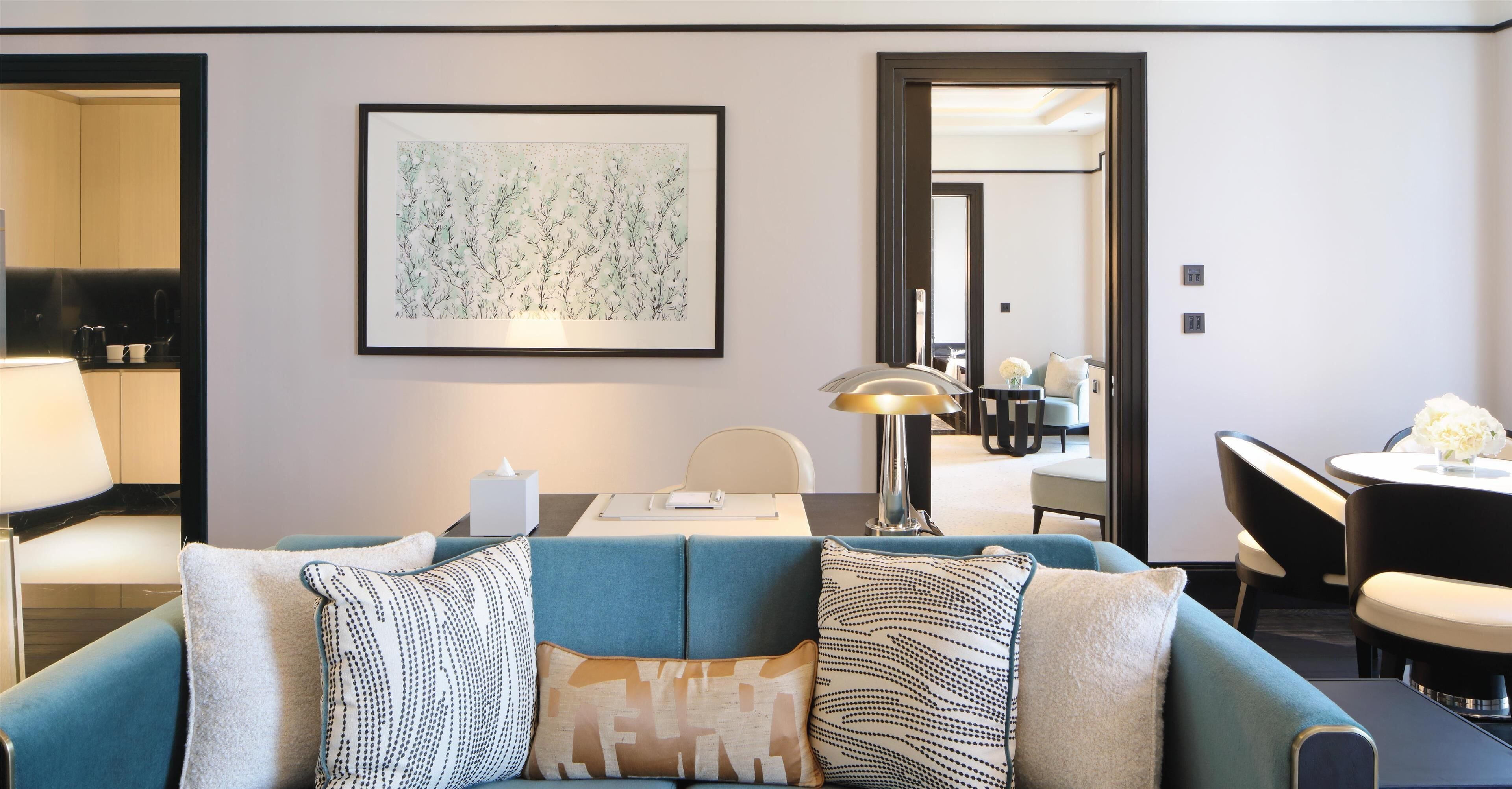 Een moderne hotelkamer met een blauwe bank, een ingelijst kunstwerk, een zithoek en een keuken die door een open deur zichtbaar is.