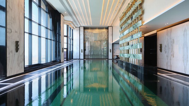 Una amplia piscina interior con superficie de agua cristalina, rodeada de paredes de mármol y grandes ventanales que dejan entrar luz natural.