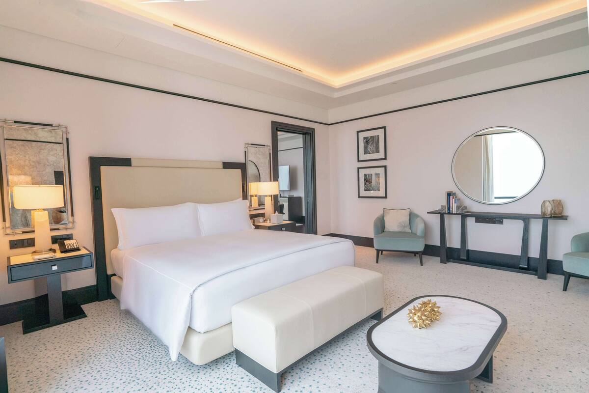 Une chambre d’hôtel moderne avec un grand lit, une table de chevet avec une lampe, un miroir rond, une petite table ronde agrémentée d’un objet décoratif et un banc.