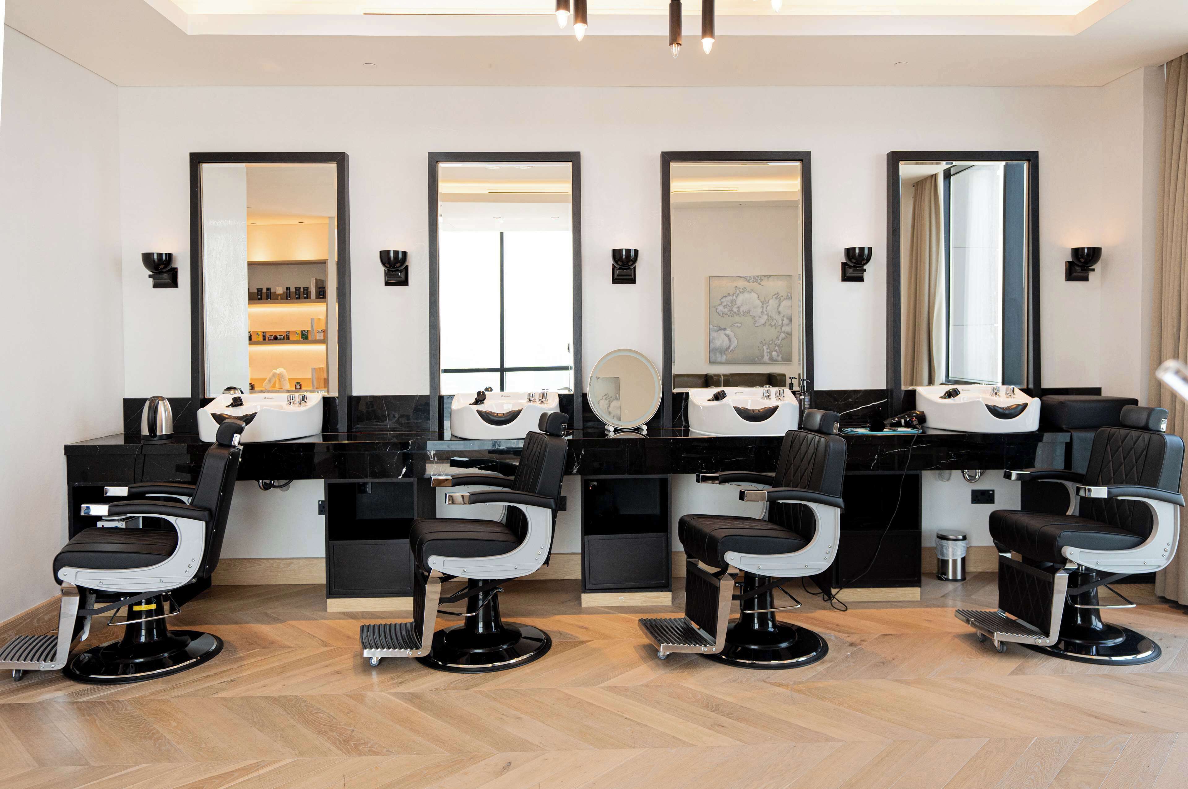 Een moderne salon met vier zwart-wit kappersstoelen, grote spiegels en een zwart aanrecht met kappersapparatuur.