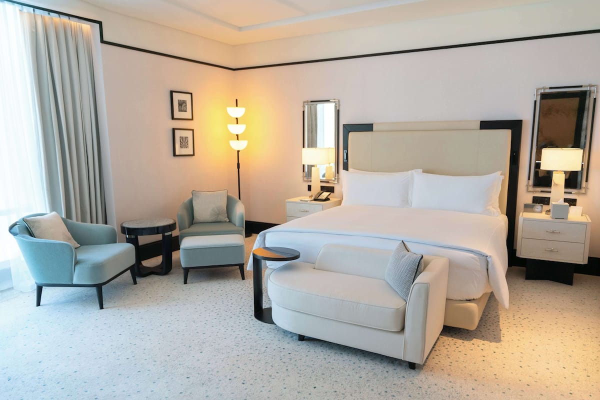 Une chambre d’hôtel avec un grand lit, deux fauteuils, une petite table, une table de chevet avec une lampe et un miroir.
