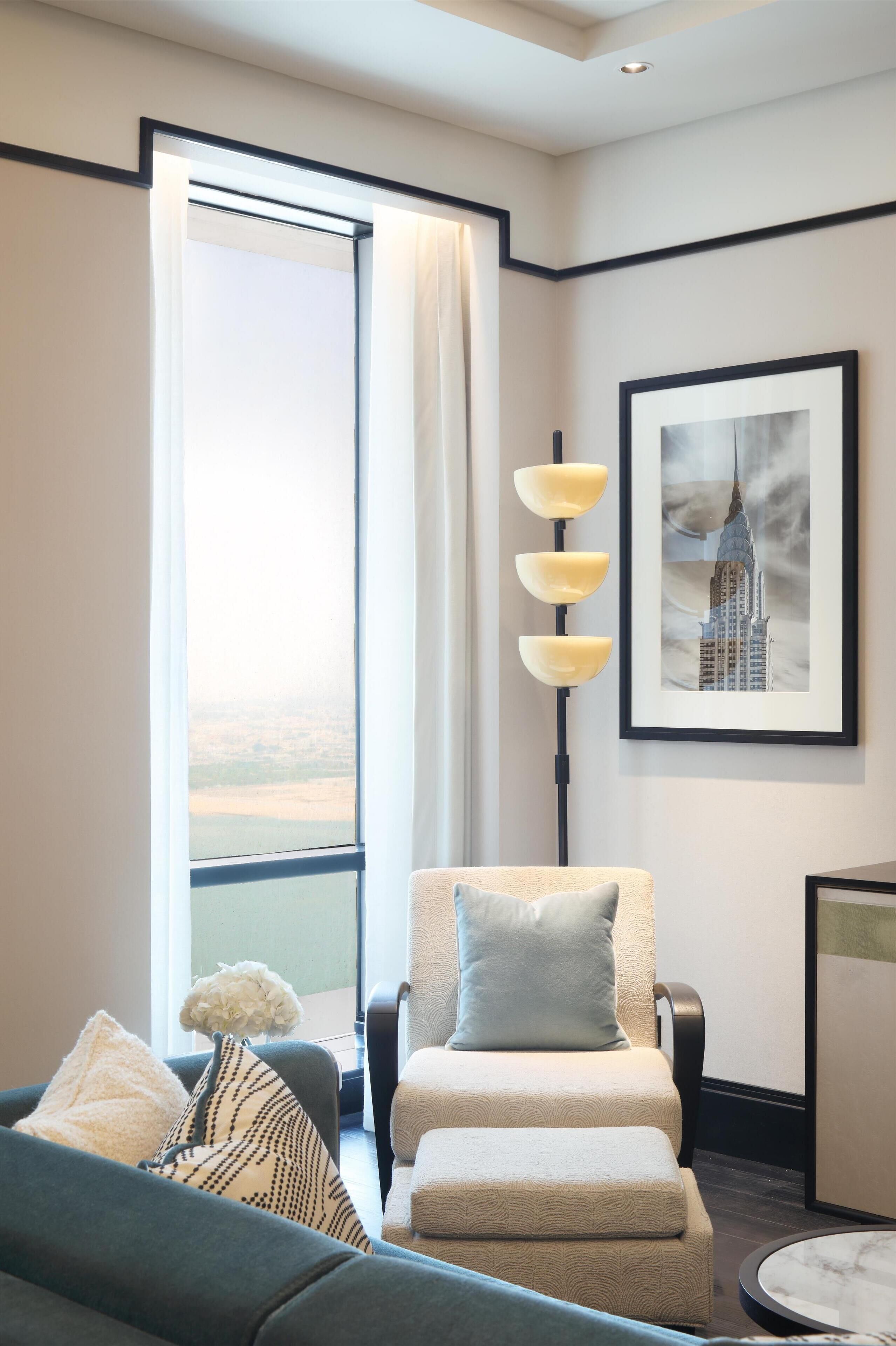 Una habitación de hotel moderna con un ventanal grande, una zona de estar acogedora con sofá y sillón, una lámpara de pie y un cuadro en la pared.