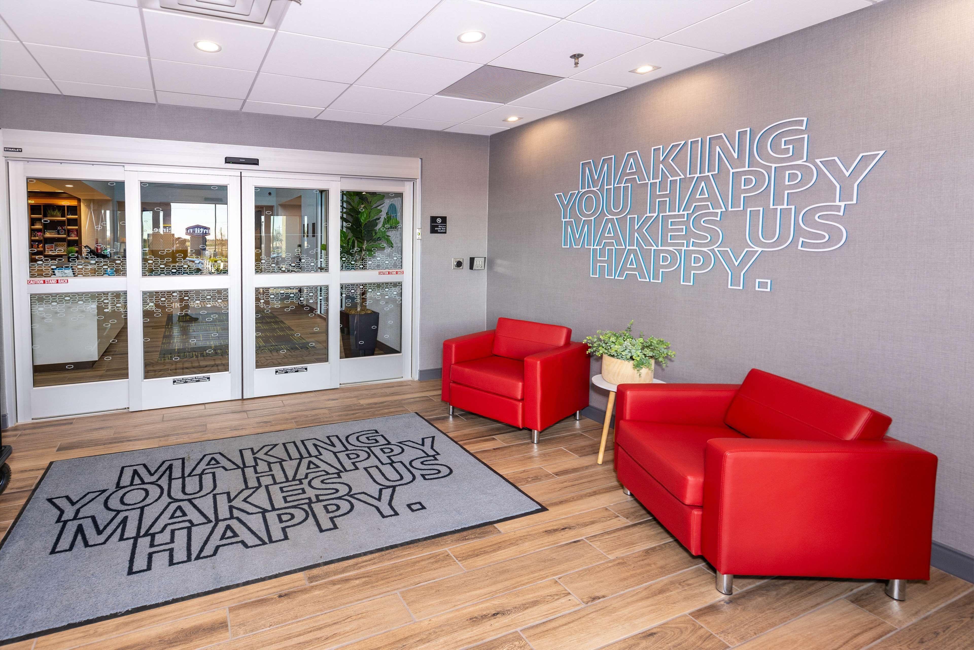 A reception area with a glass door entrance, a red sofa, a small table with a plant, and a welcome mat with the text "MAKING YOU HAPPY MAKES US HAPPY."