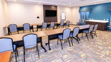 A conference room with a long table, chairs, a flat-screen TV, a whiteboard, and a kitchen area with a counter and sink.