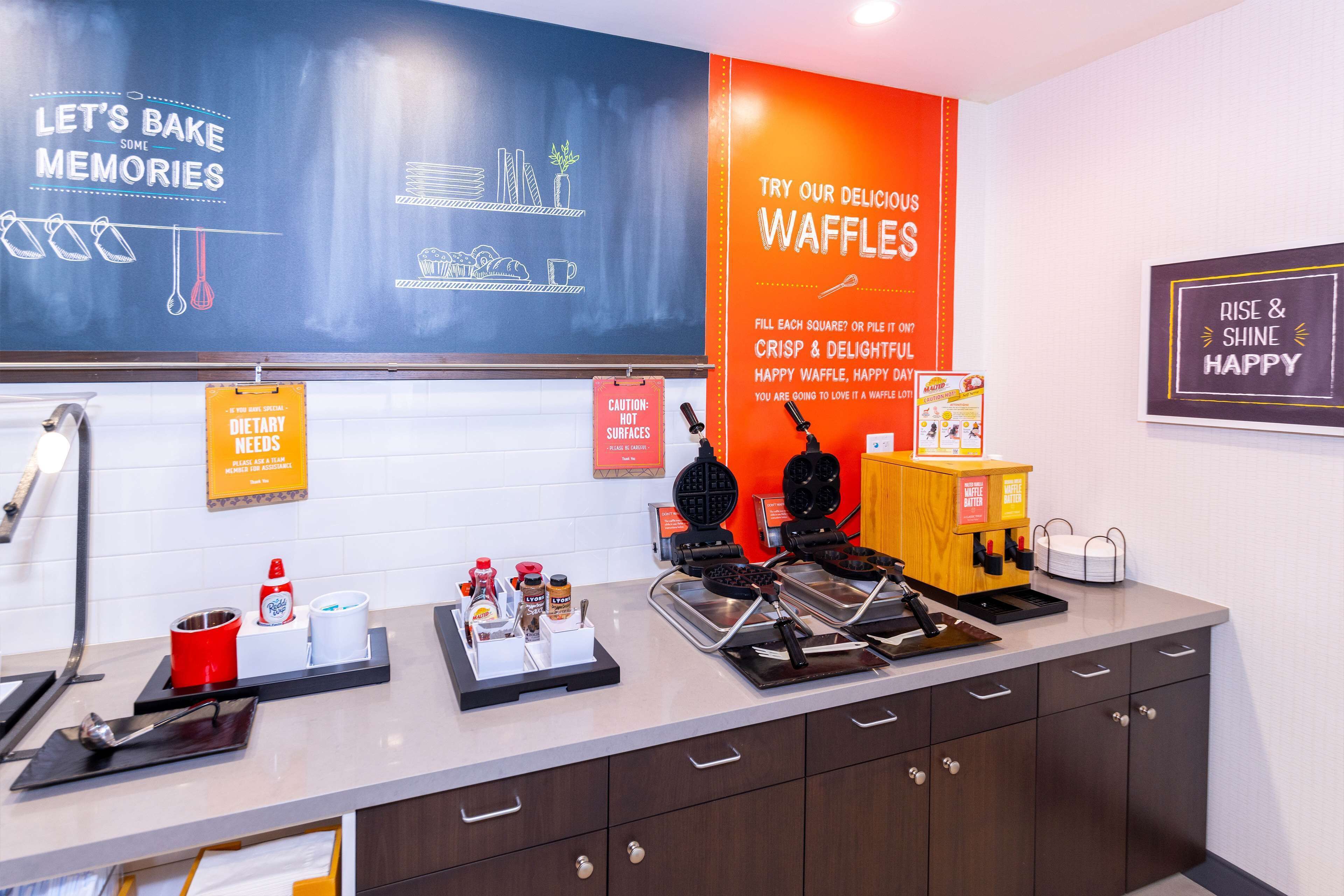 A kitchen area with waffle-making equipment, a chalkboard with baking tips, and a sign promoting waffles.