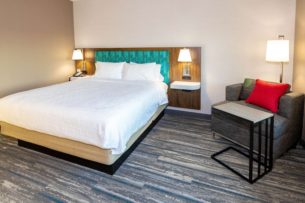 Ein Hotelzimmer mit einem großen Bett, zwei Nachttischen mit Lampen, einem kleinen Sofa mit rotem Kissen und einem gemusterten Teppich.