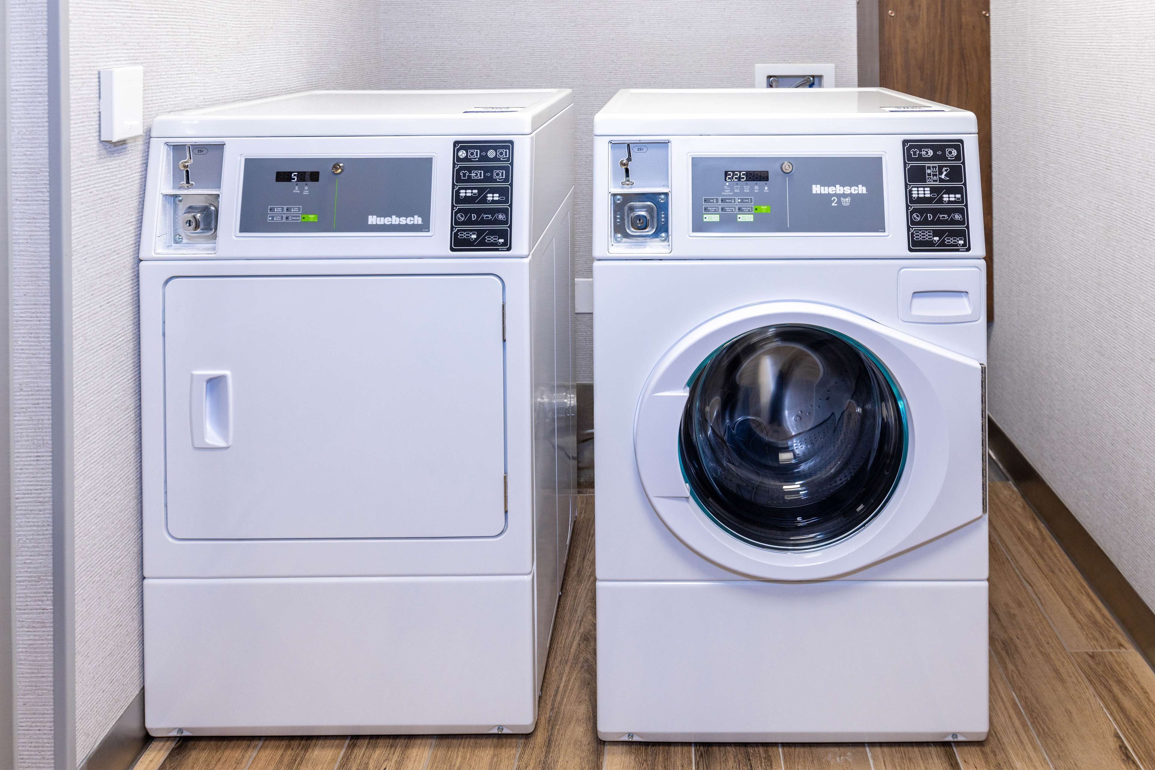 Two white front-loading washing machines side by side in a laundry room with a wooden floor and a light-colored wall.