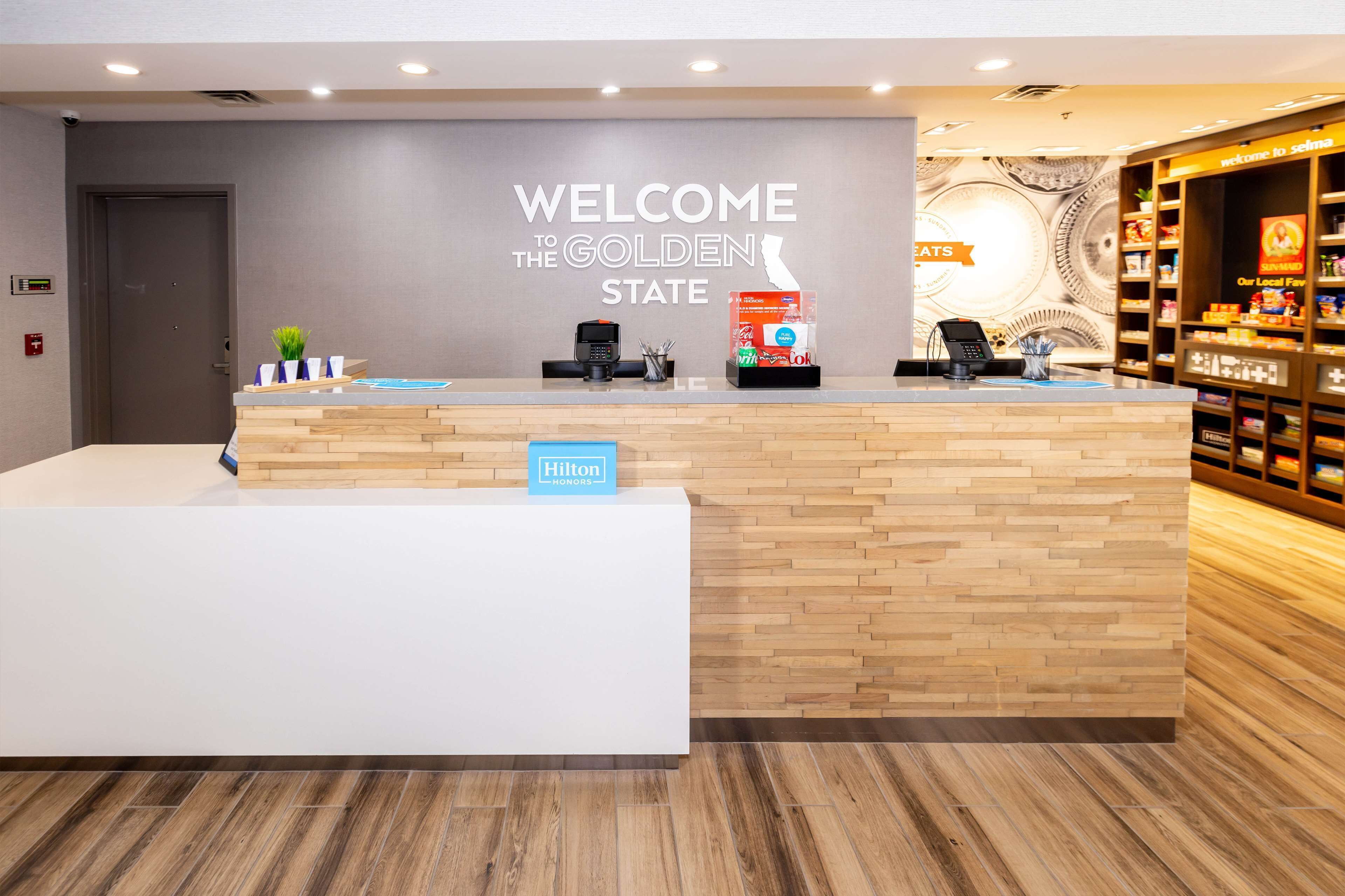 A hotel reception desk with a "Welcome to the Golden State" sign, a "Hilton Honors" sign, and a counter with a coffee machine and a phone.