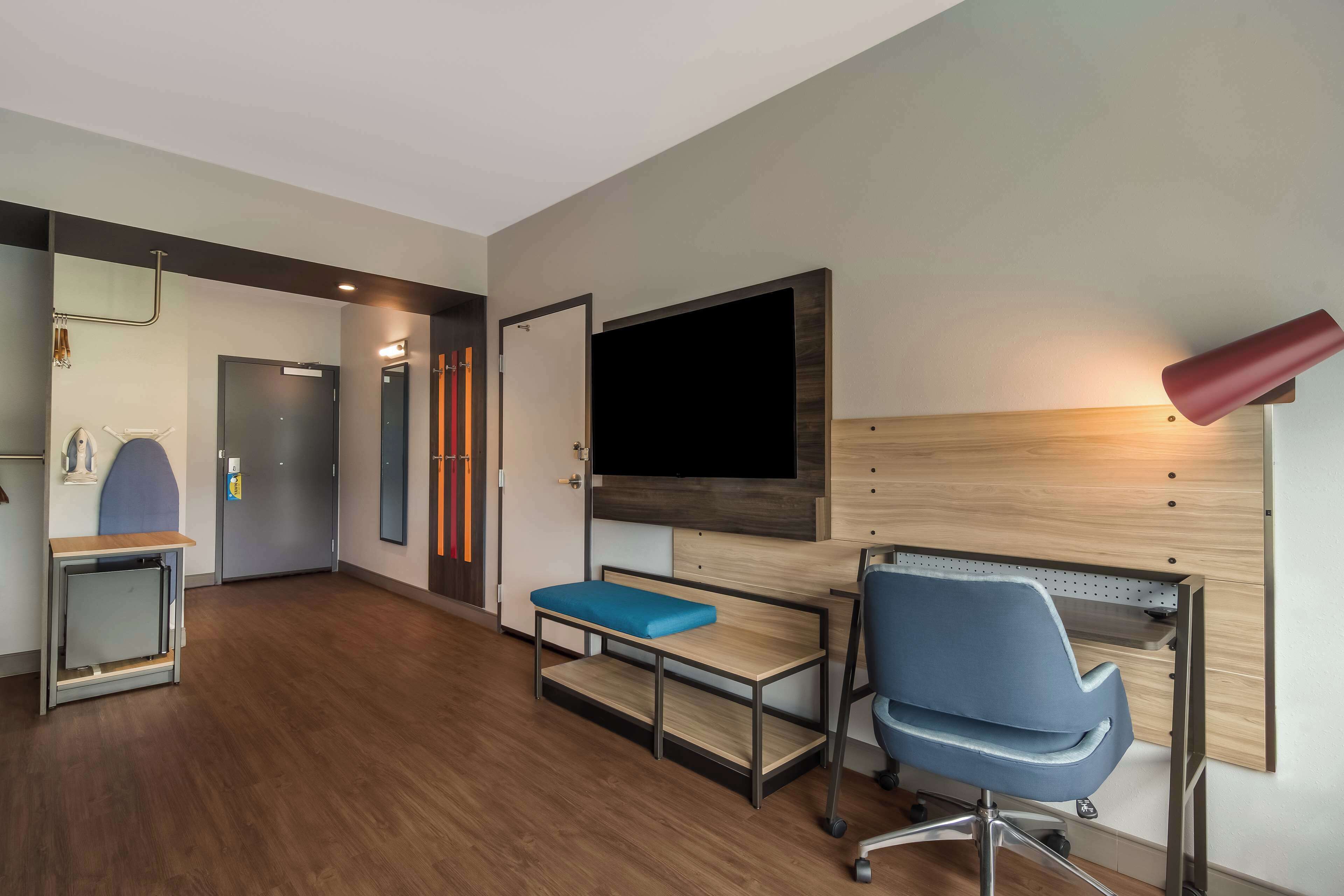 Una habitación de hotel moderna con piso de madera, un escritorio con una silla azul, un televisor de pantalla plana montado en la pared y un banco con un cojín azul.