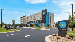 Een modern hotel met een opvallend 'tru by Hilton' bord, een parkeerplaats en een heldere lucht.