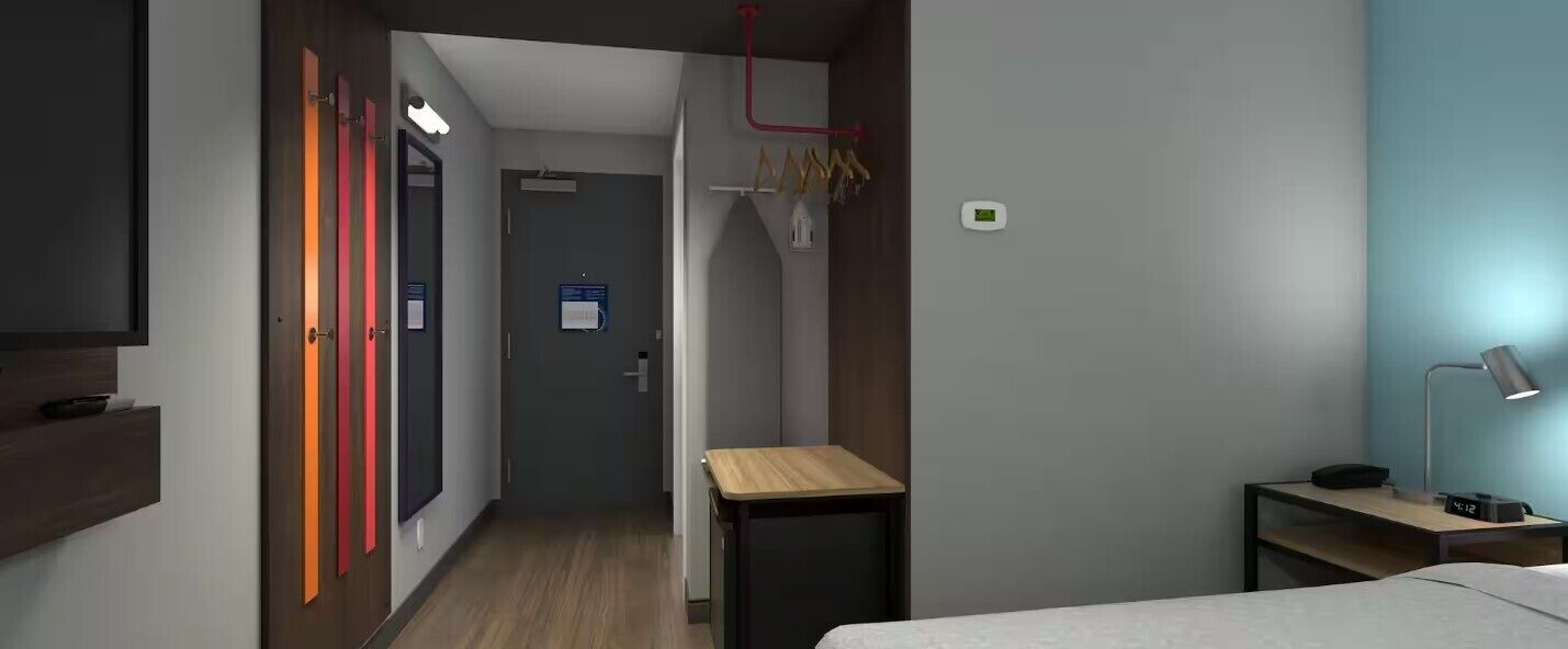 Habitación de hotel con cama, mesita de noche, lámpara y un escritorio pequeño con silla. Se ve un armario con espejo y una puerta con pomos de colores. En la esquina, un perchero.