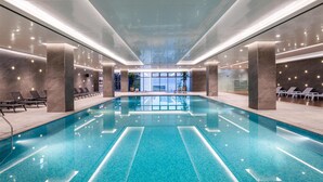 Una piscina interior moderna con agua cristalina, rodeada de sillones de descanso y un muro de vidrio con vistas a la ciudad.
