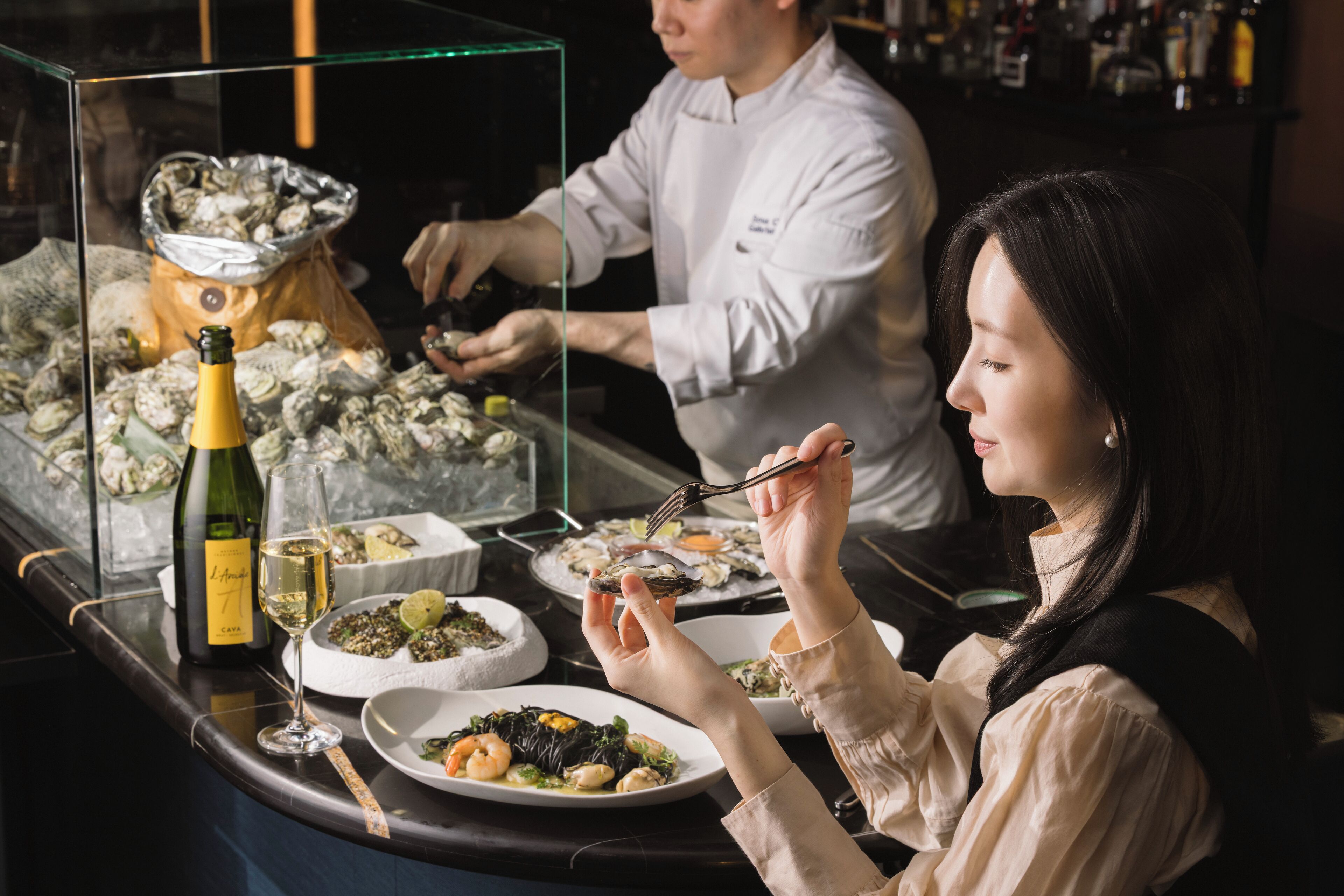 A dining area with a chef preparing food and a guest dining at a table with various dishes and a bottle of wine.