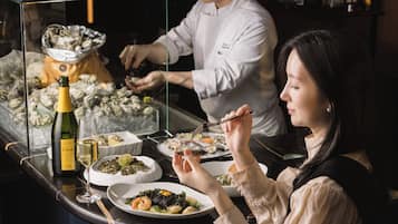 A dining area with a chef preparing food and a guest dining at a table with various dishes and a bottle of wine.