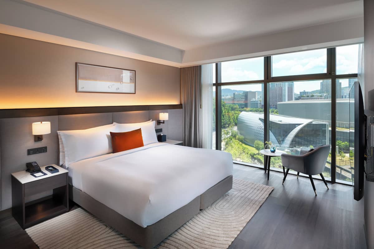 A modern hotel room with a large bed, bedside lamps, a small table, a chair, and a large window offering a city view.