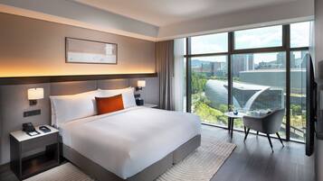 A modern hotel room with a large bed, bedside lamps, a small table, a chair, and a large window offering a city view.