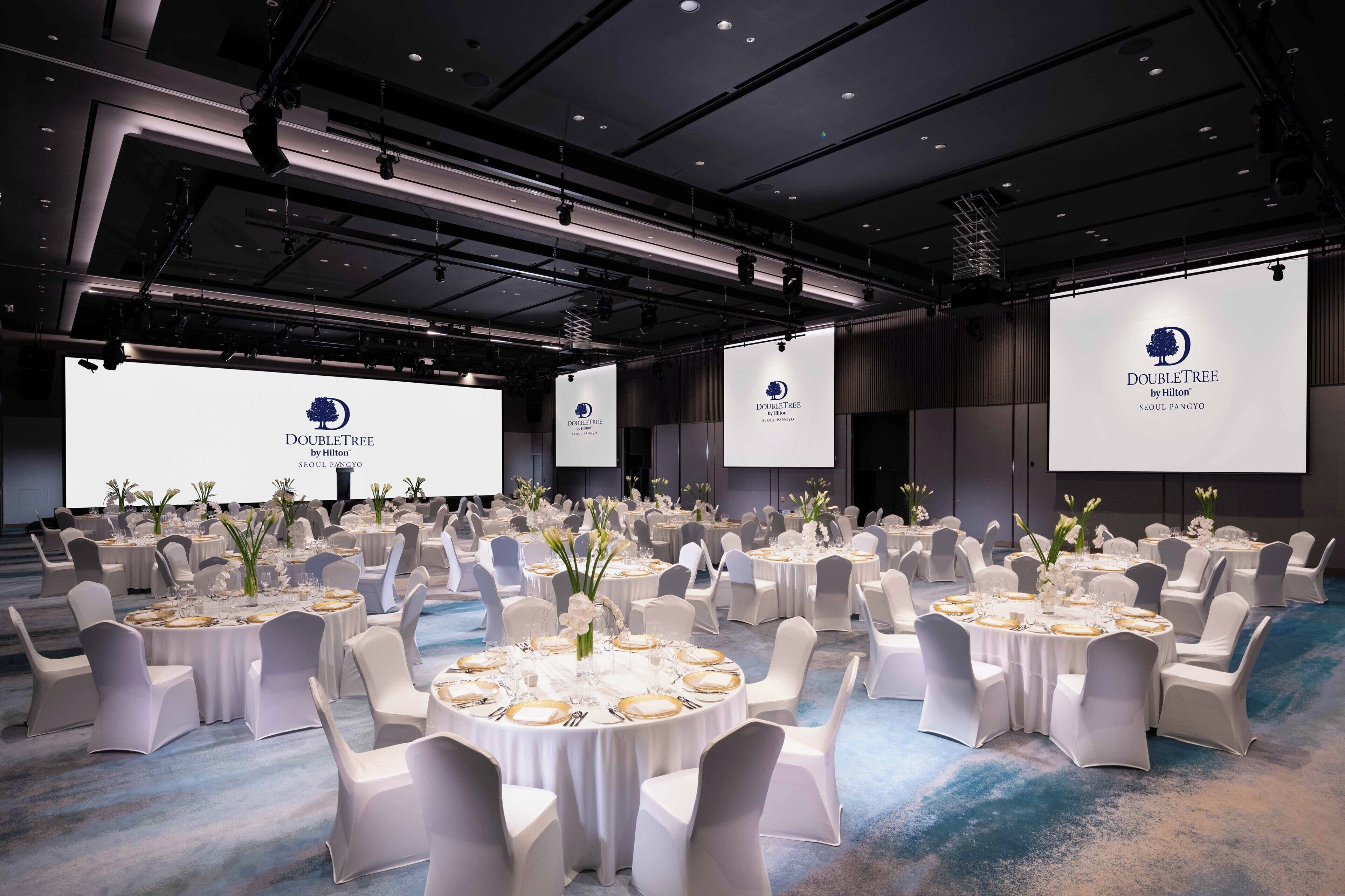 A large conference room with multiple round tables set for events, each with white chairs and floral centerpieces.