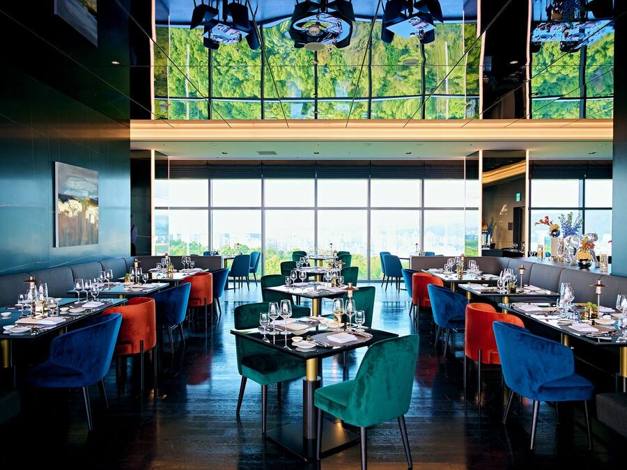 A modern restaurant with high ceilings, large windows offering a city view, and vibrant seating arrangements.
