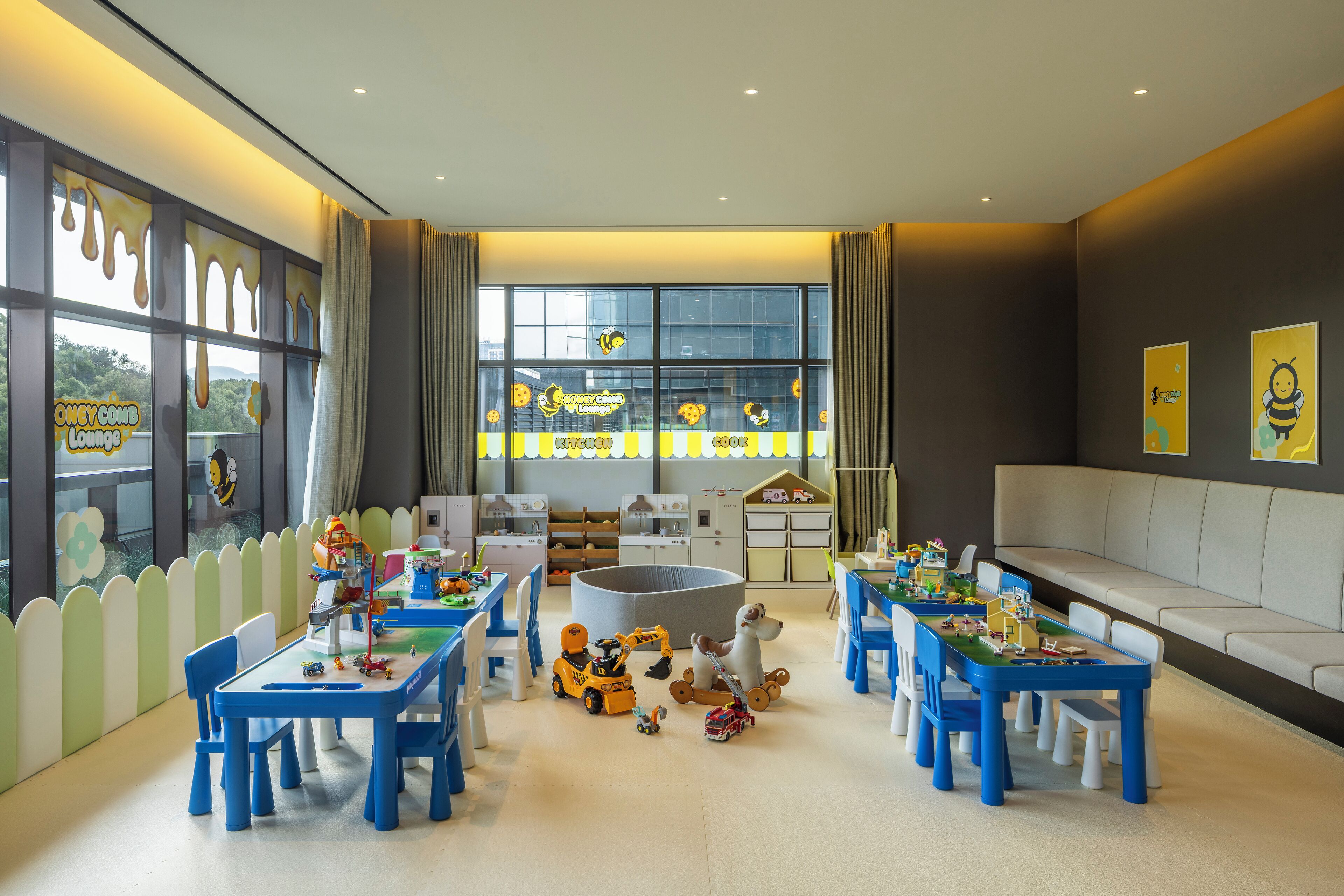 A children's playroom with multiple tables and chairs, a large window with a view, and a play area with toys and a slide.