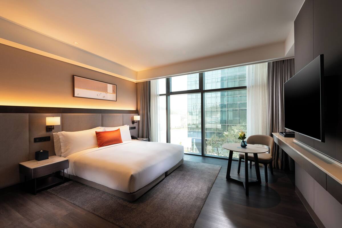 A hotel room with a large bed, bedside tables, a flat-screen TV, a small table with chairs, and a large window offering a city view.