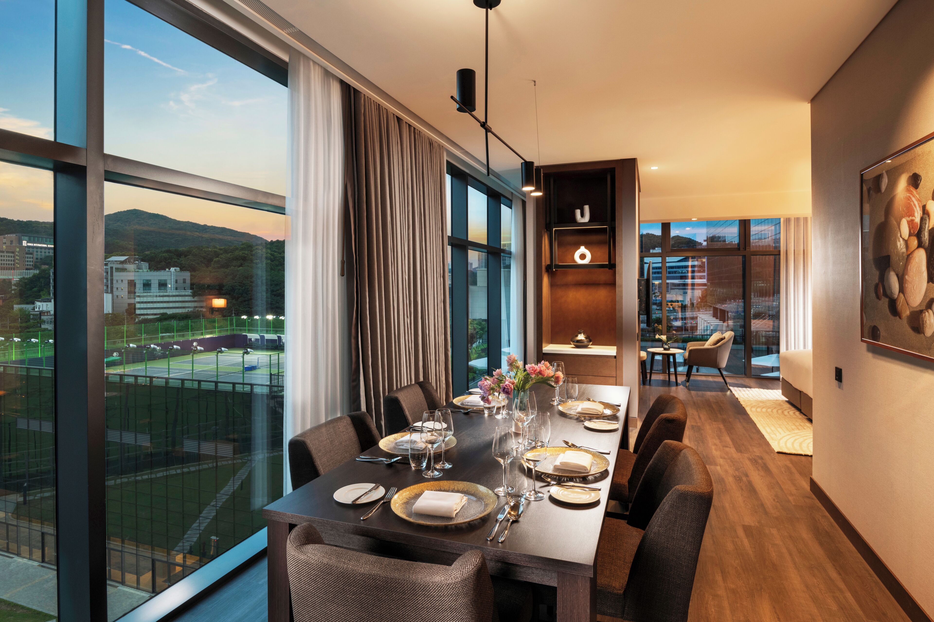 Un comedor con una mesa larga dispuesta para seis personas, ventanas amplias y vista a un campo deportivo.