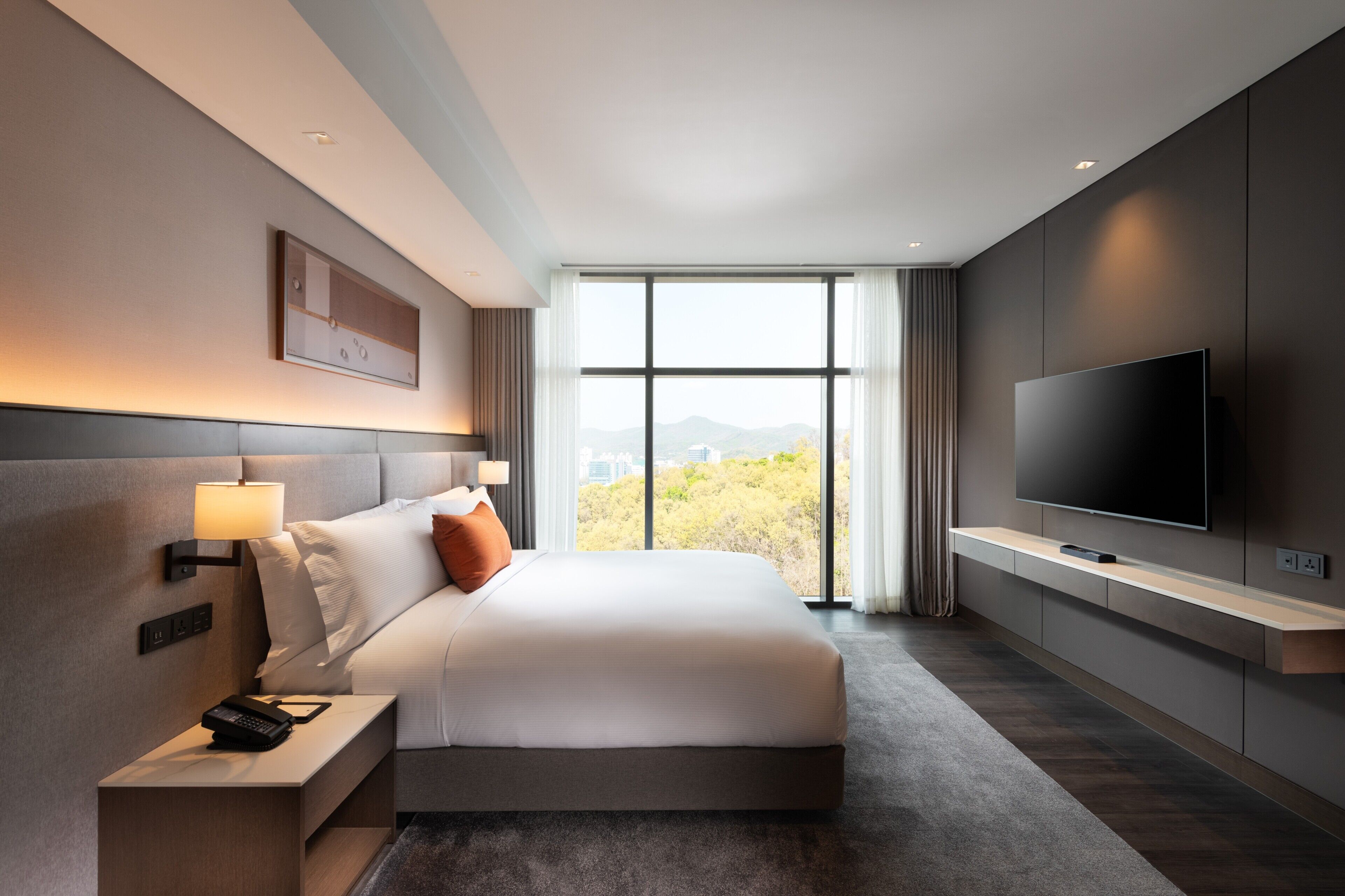 A modern hotel room with a large bed, a TV, a nightstand, and a window with a view.