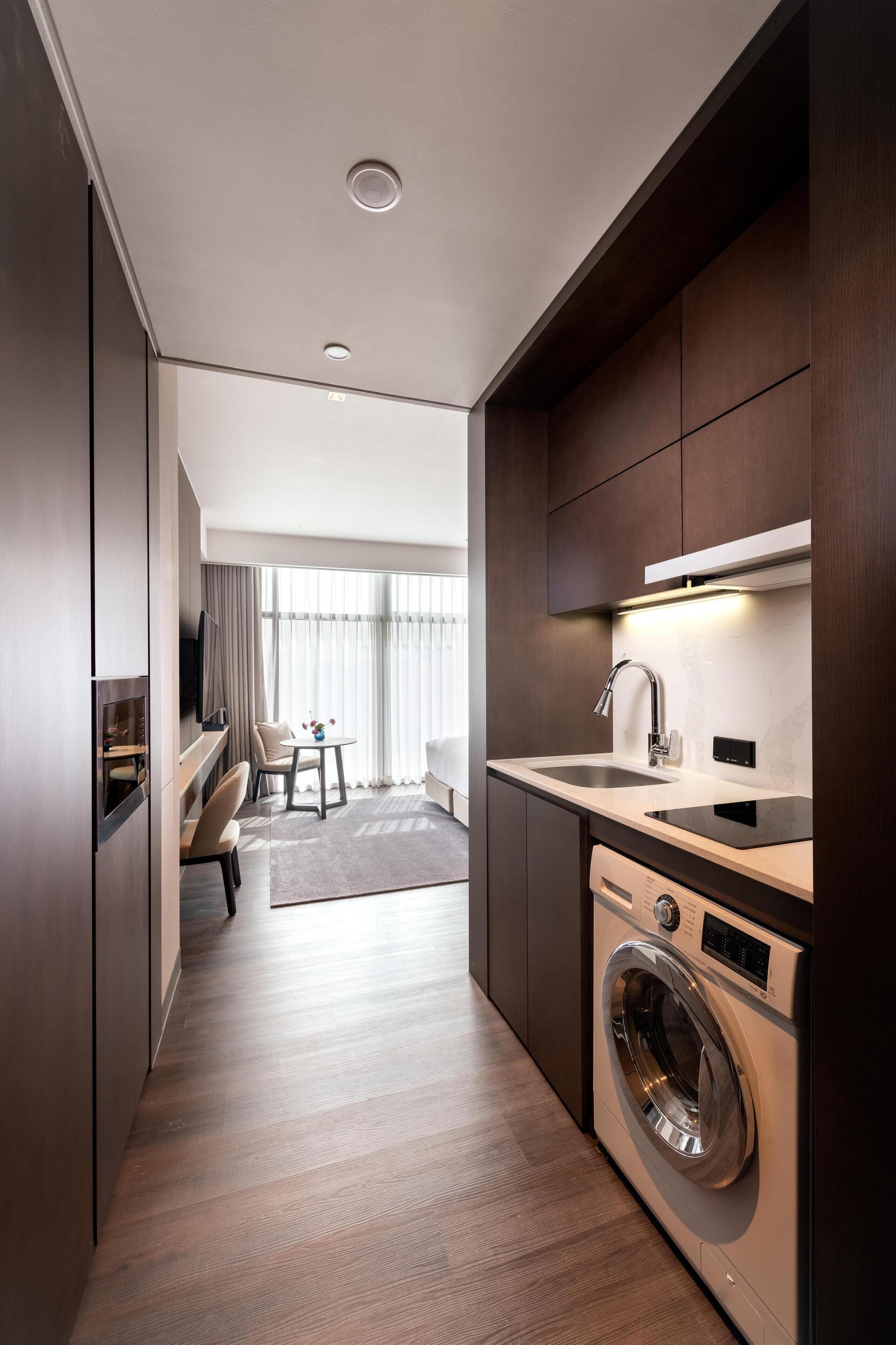 A modern hotel room with a compact layout, including a kitchenette, a washing machine, a seating area with a sofa and chairs, and a large window providing natural light.