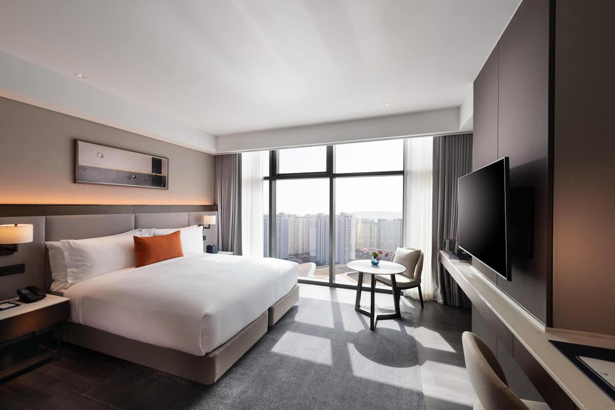 A modern hotel room with a large bed, a TV, a desk with a lamp, a small table, and a view of the city.