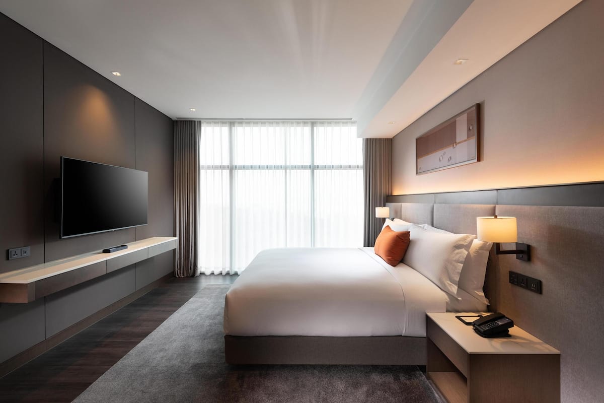 A modern hotel room with a large flat-screen TV mounted on the wall, a bed with white bedding, a bedside table with a lamp, and a large window with sheer curtains.