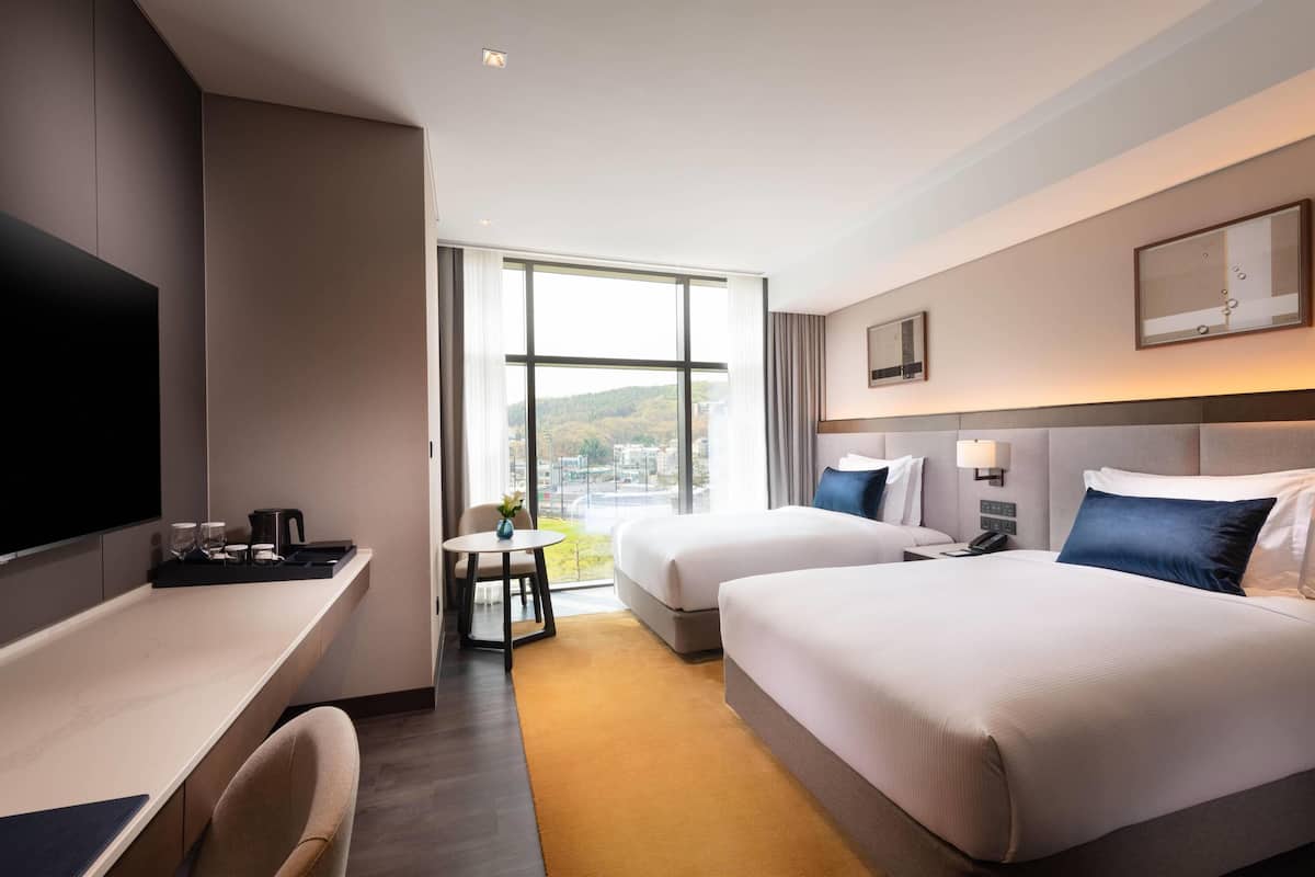 A hotel room with two beds, a desk, a TV, and a large window offering a city view.