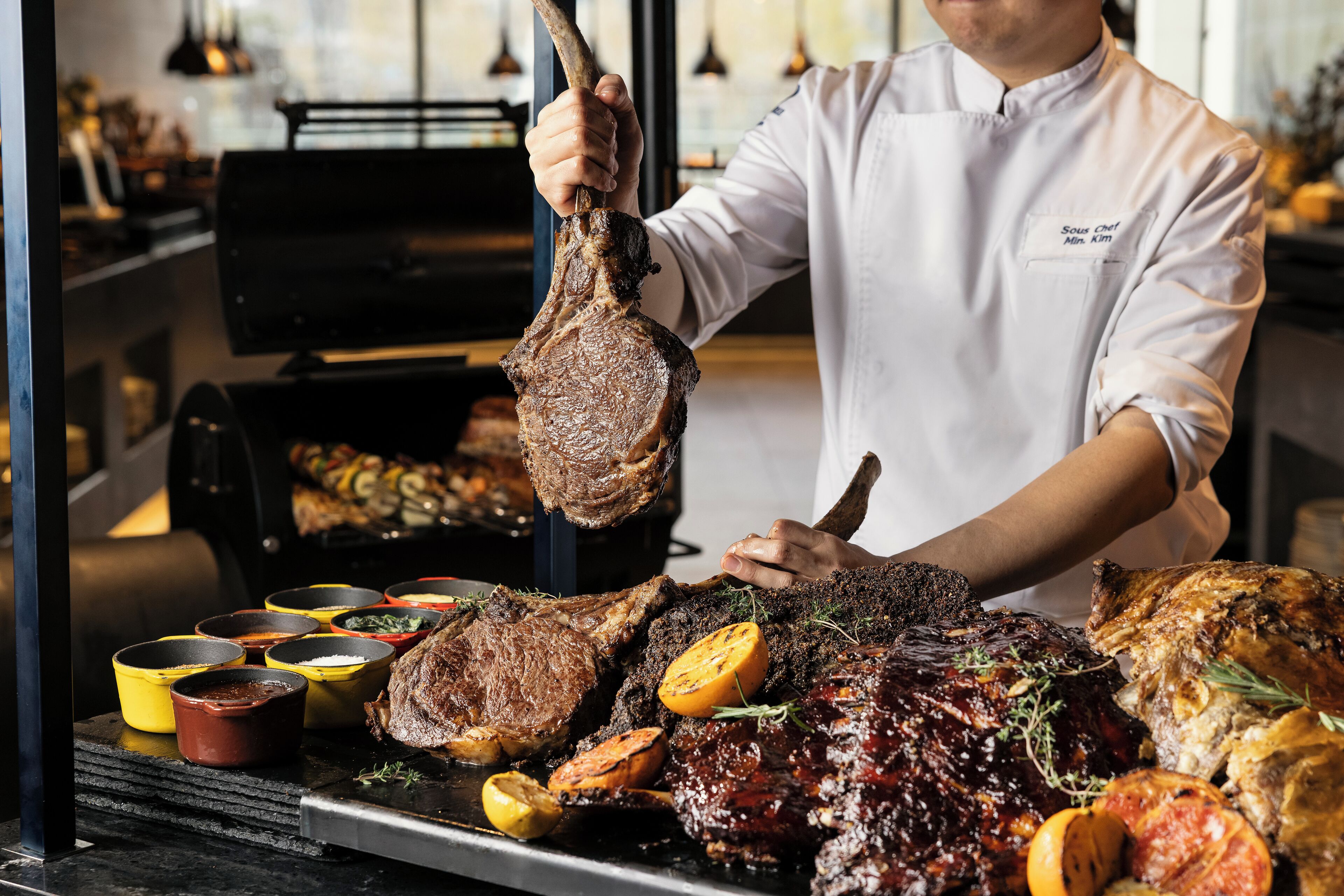 Un chef tenant un gros morceau de viande, entouré de différents plats et ustensiles de cuisine.