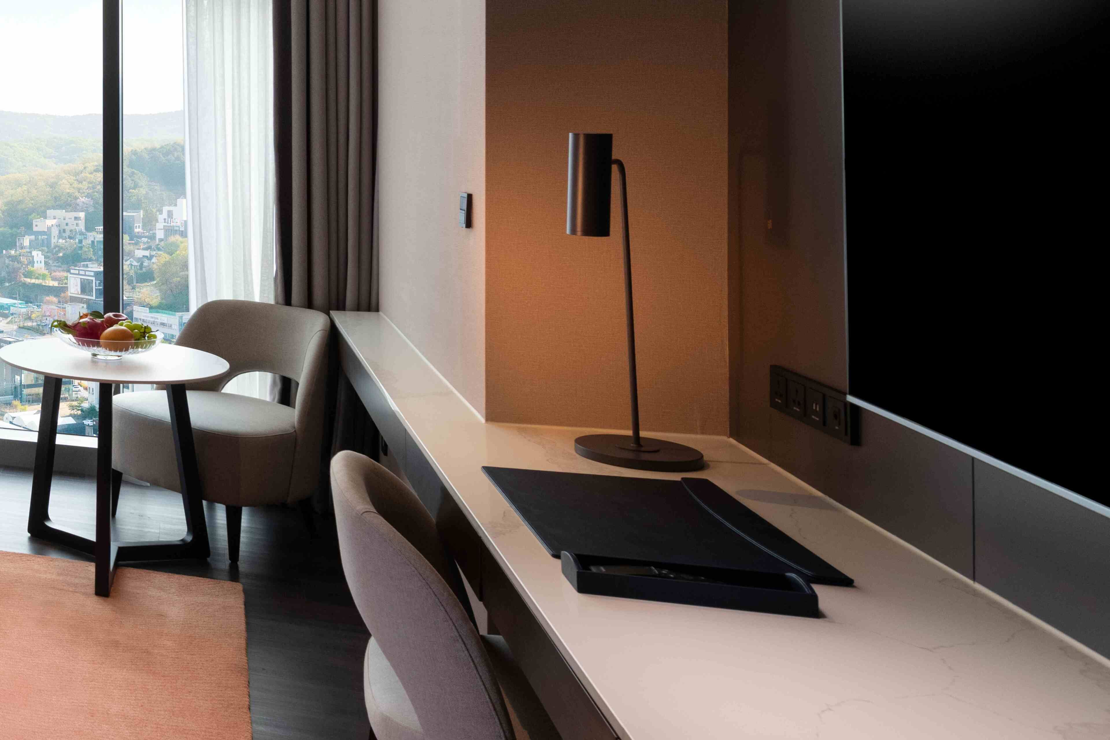 A modern hotel room with a large window, a desk with a laptop, a chair, a small table with fruit, and a floor lamp.