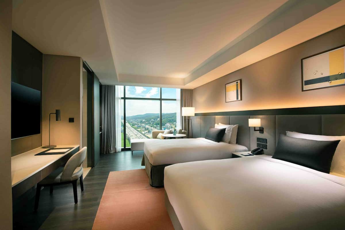 Habitación de hotel con dos camas, un escritorio, televisor y un ventanal con vistas a la ciudad.