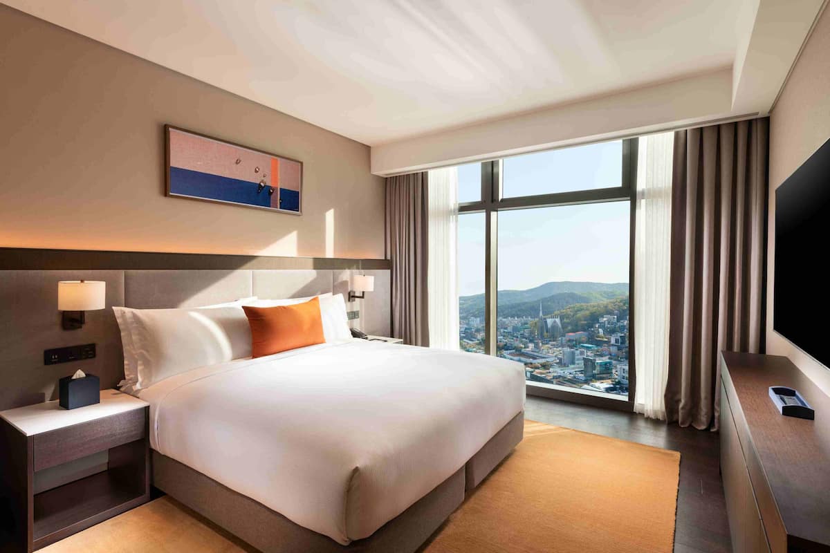 Habitación de hotel con una cama grande, una mesita de noche, un televisor y una ventana con vistas a la ciudad.