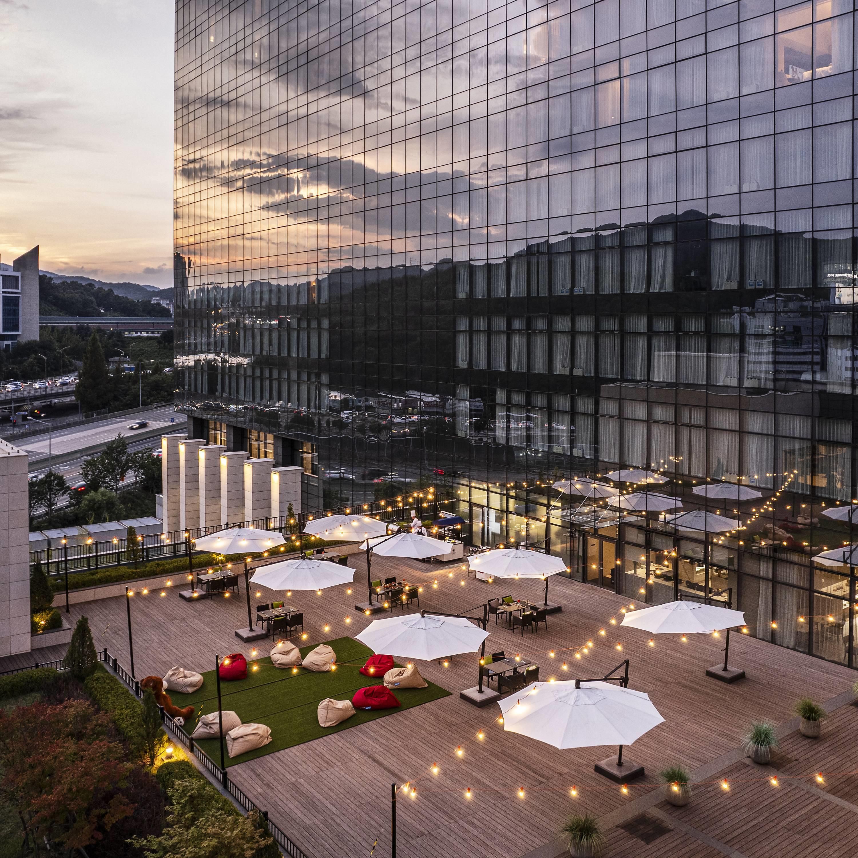 A rooftop terrace with outdoor seating and umbrellas, surrounded by a modern building with reflective glass windows.