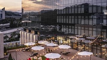 A rooftop terrace with outdoor seating and umbrellas, surrounded by a modern building with reflective glass windows.