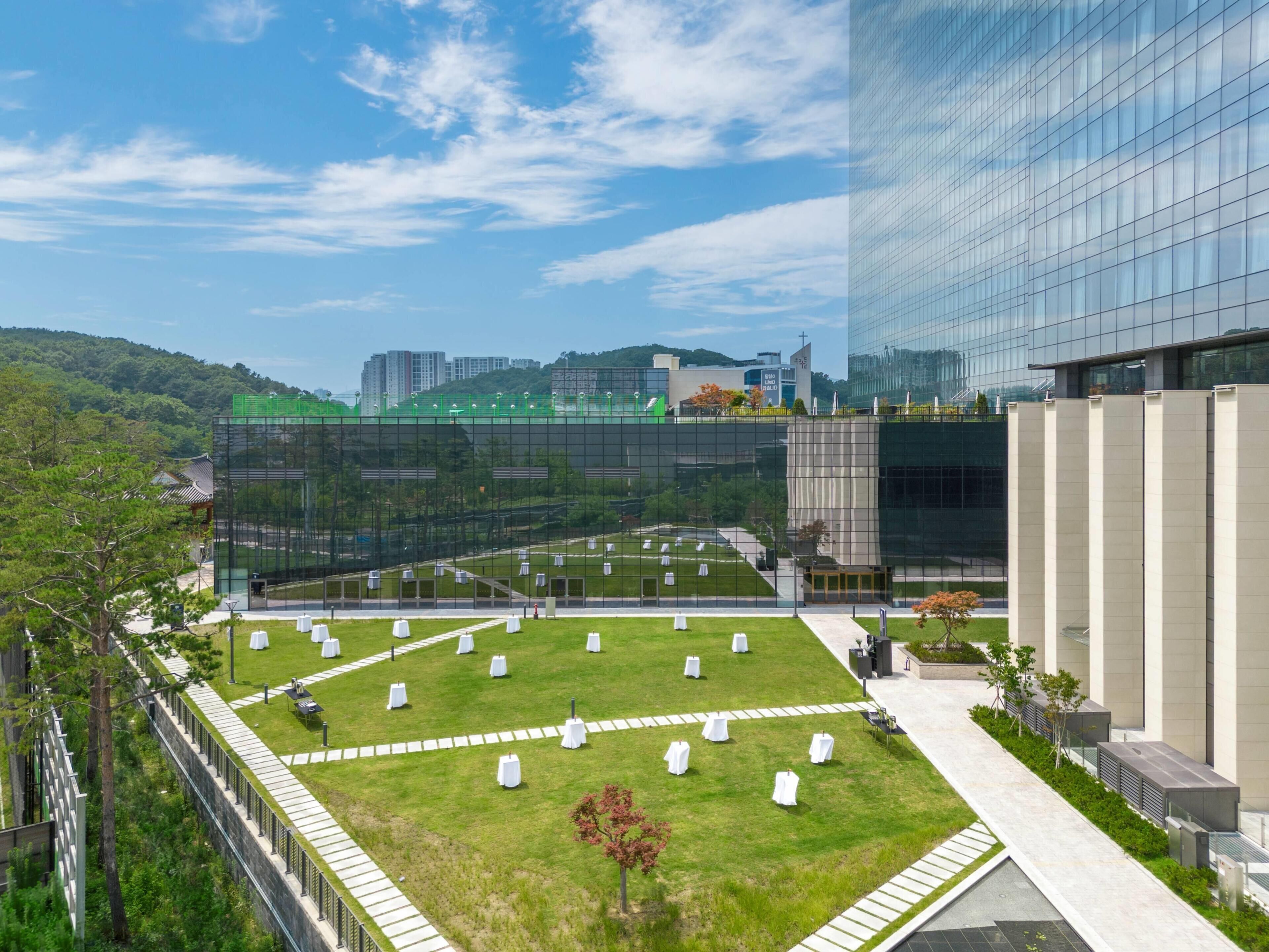 A modern building with a glass facade, a grassy courtyard with white tables, and a clear blue sky.