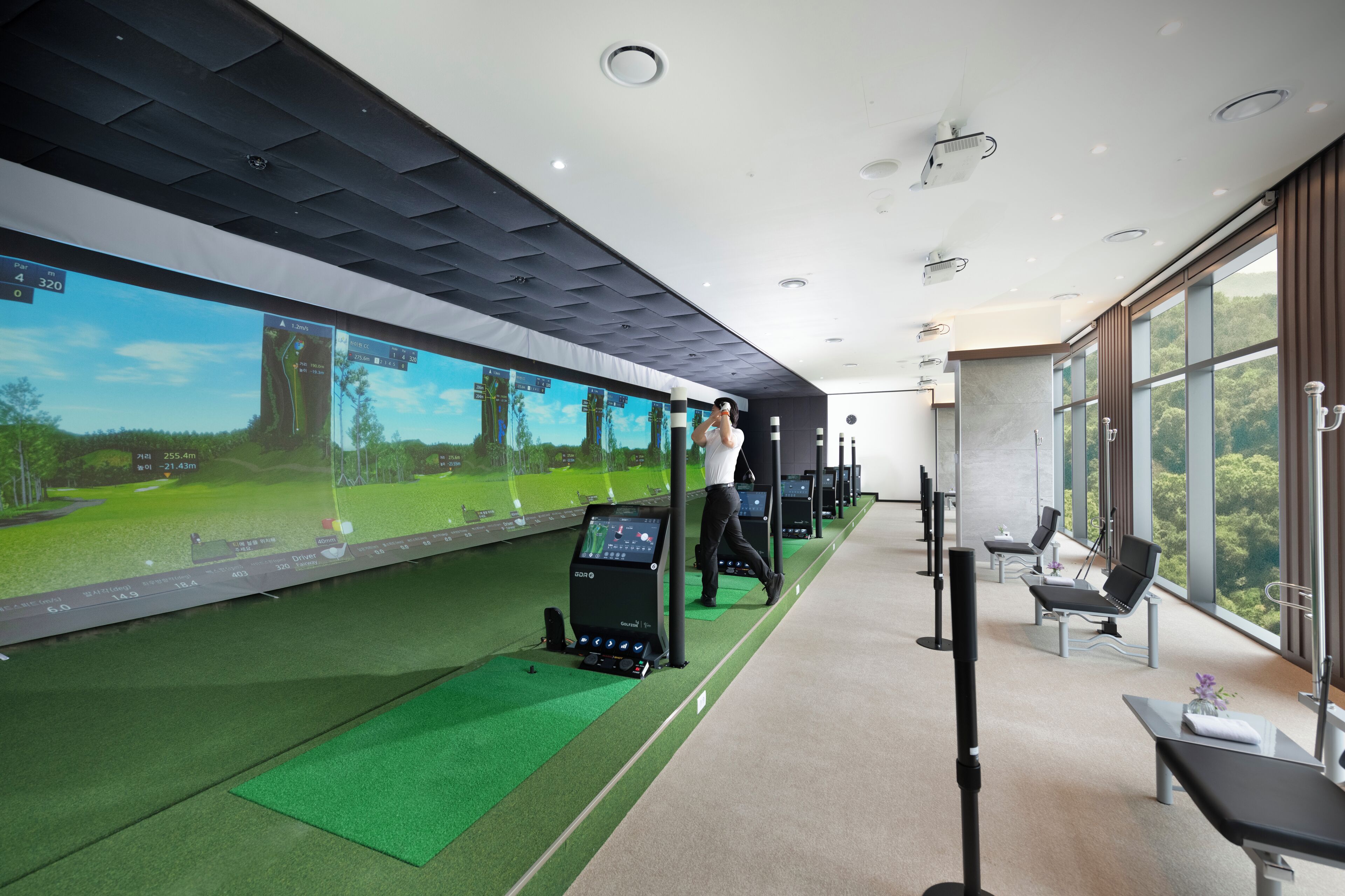 A modern indoor golf simulation room with a large screen displaying a golf course, a golf cart, and a control panel.