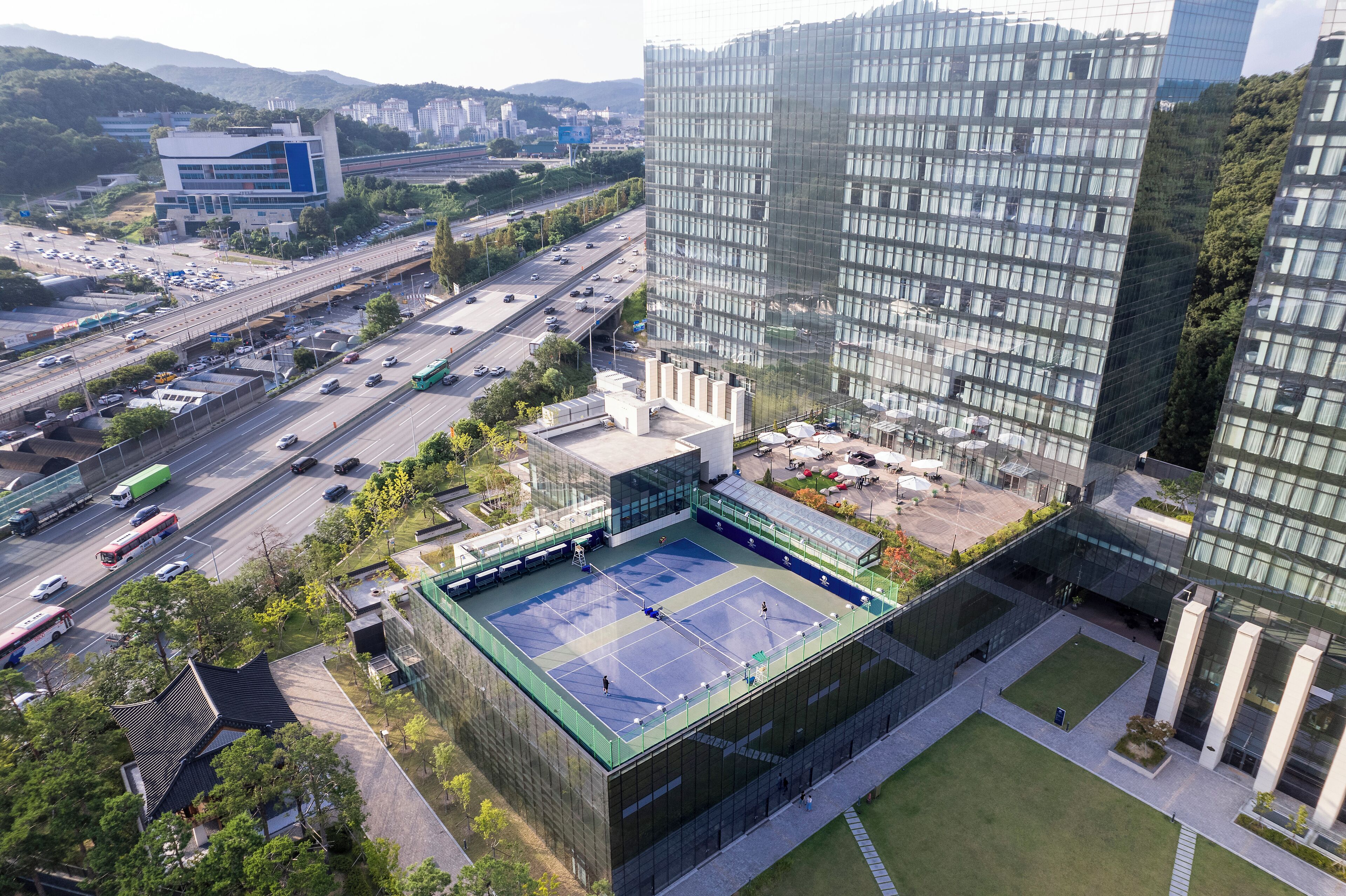 Um edifício moderno com quadra de tênis no terraço, cercado por infraestrutura urbana e vegetação.