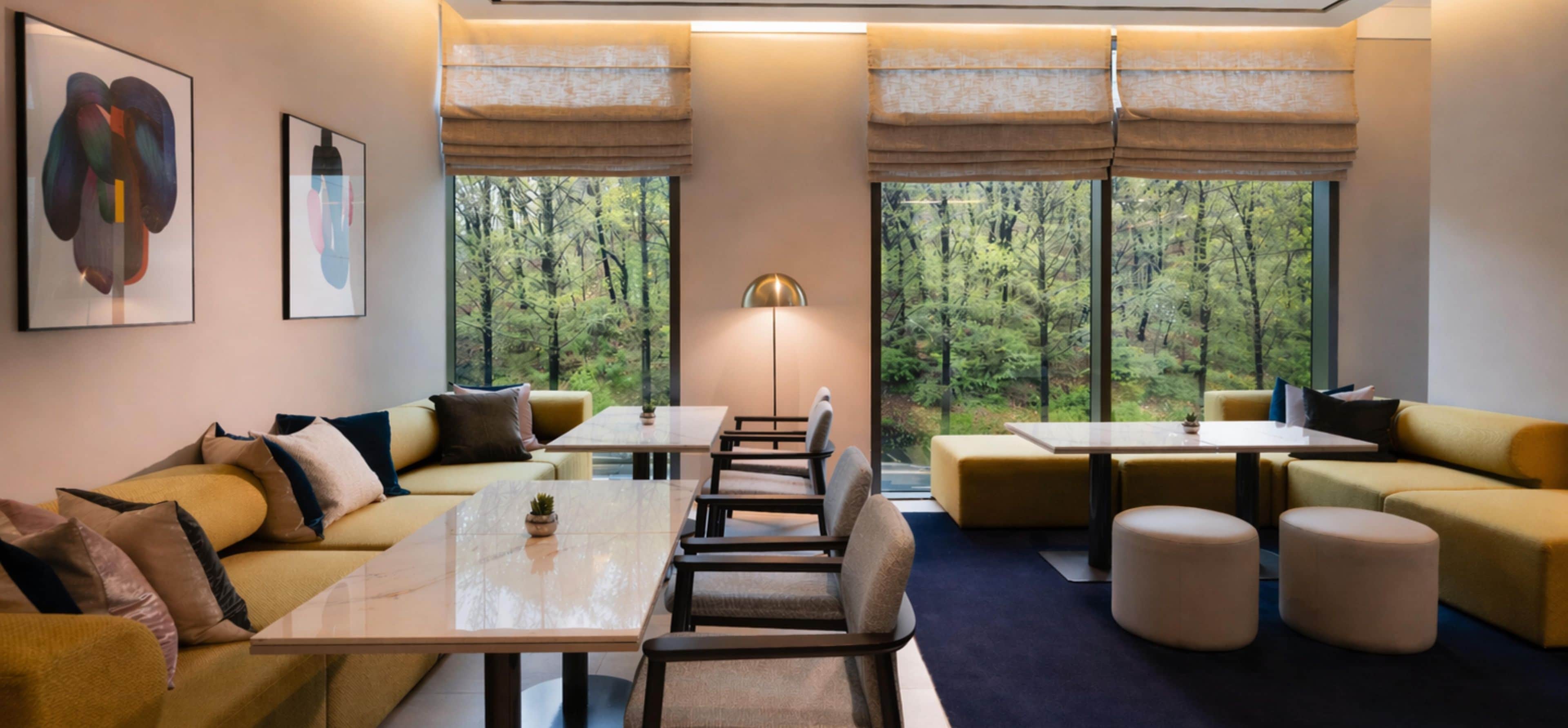 A modern hotel room with a sofa, tables, and a view of a forest through large windows.
